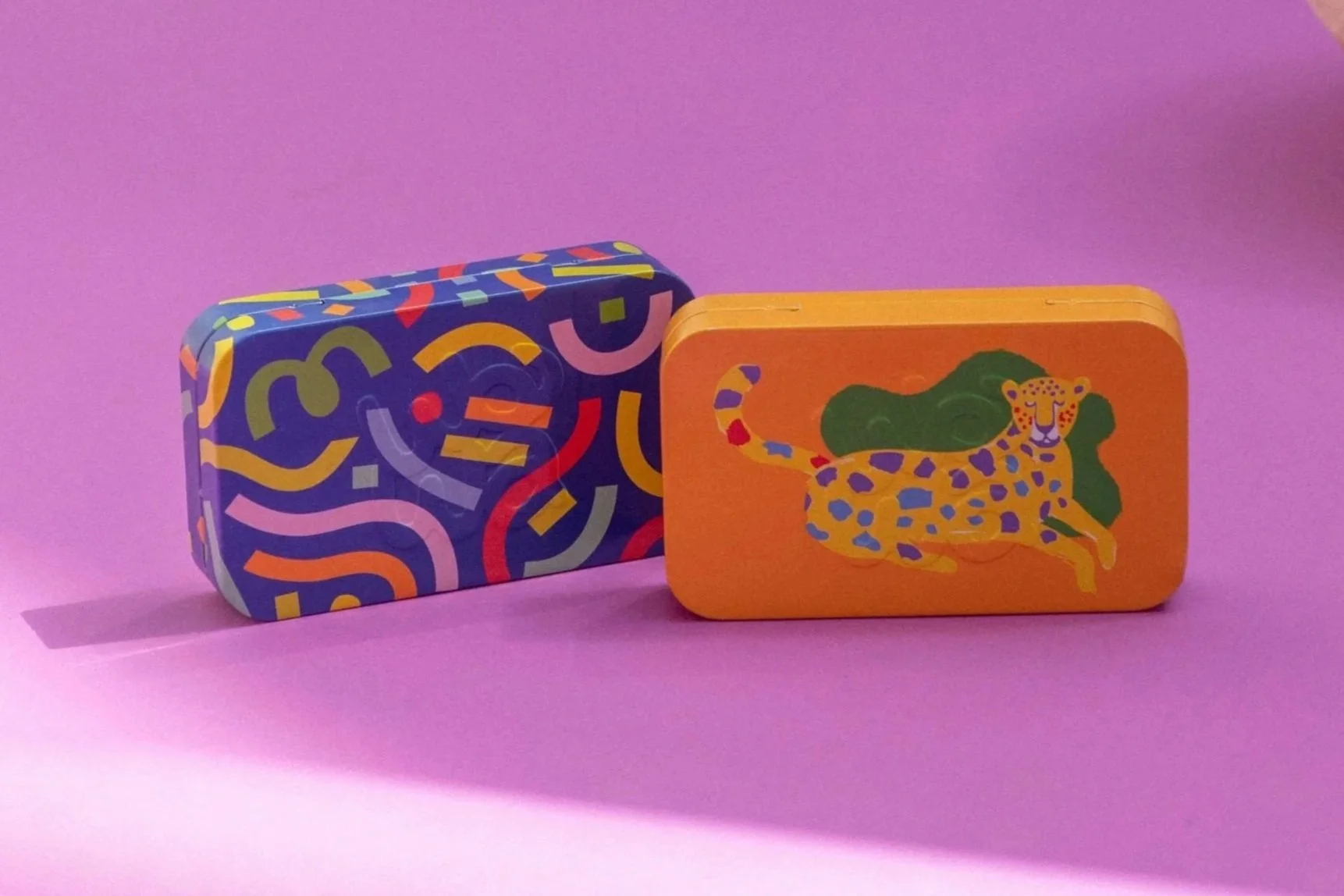 Two colorful rectangular tins with lids, one with a multicolored abstract pattern and the other with a cheerful leopard illustration on a bright orange background, positioned on a light pink surface against a light purple background.