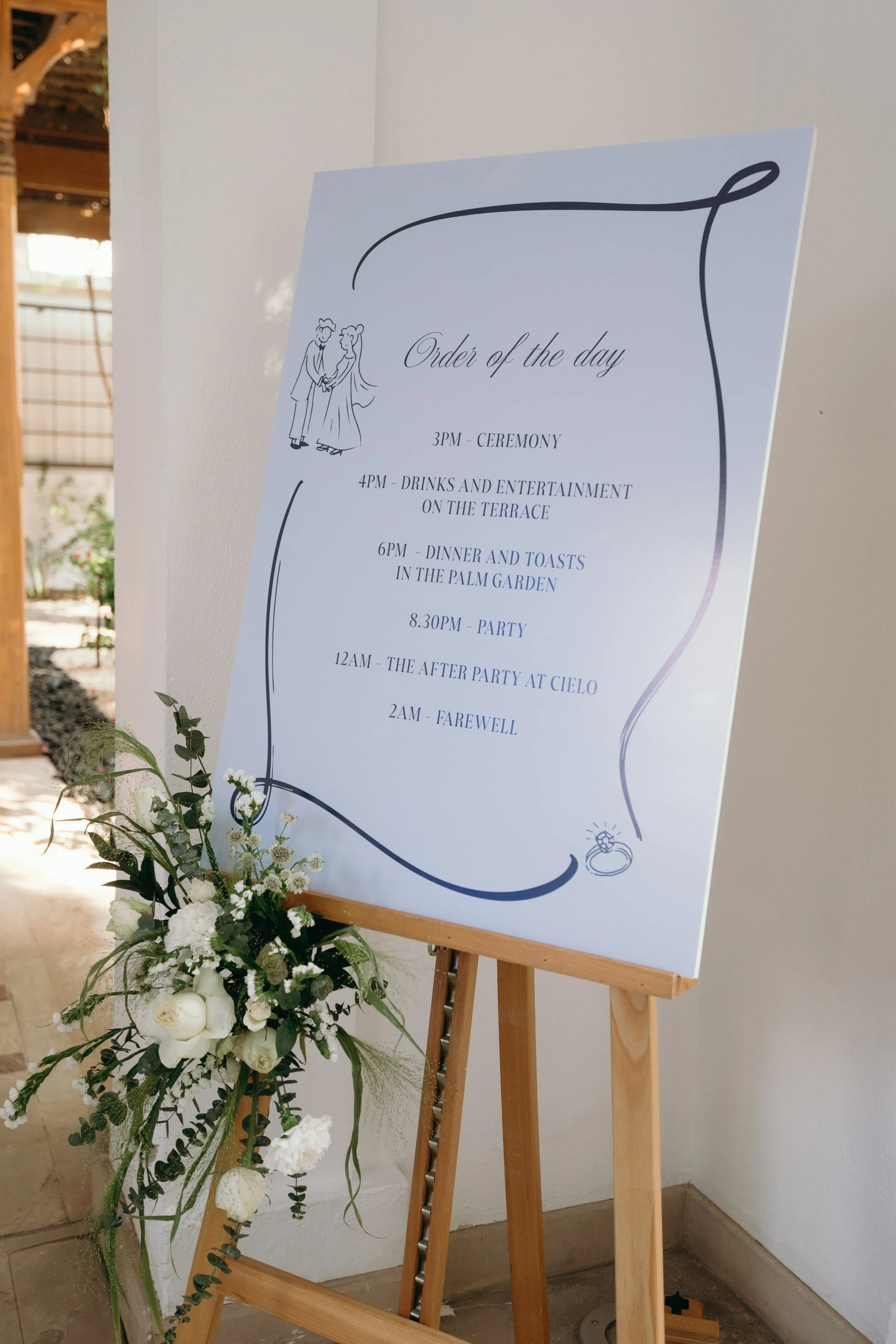 Wedding ceremony and reception schedule on a white poster board on an easel, next to a floral arrangement with white flowers and greenery, in a decorated venue with wooden elements.