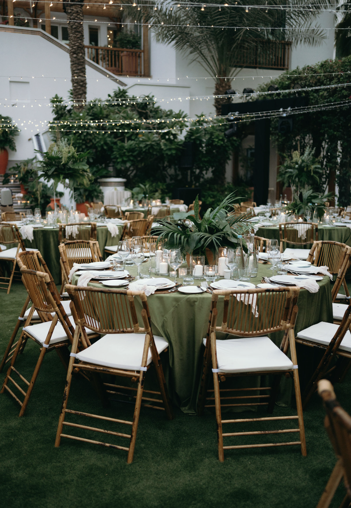 Outdoor event space with round tables covered in green tablecloths, decorated with candles and lush green plants, surrounded by wooden chairs with white cushions, under string lights, with palm trees and a white building in the background.