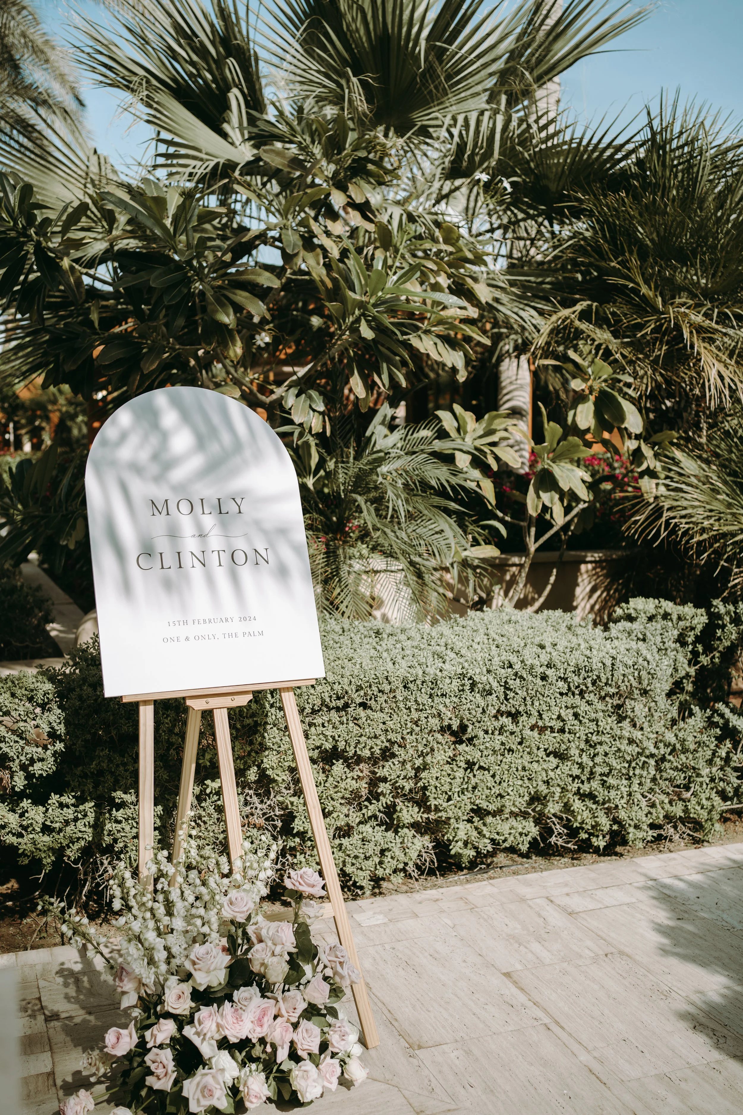 A wedding sign on an easel surrounded by pink and white roses with greenery, set against a backdrop of tropical palm trees and blue sky.