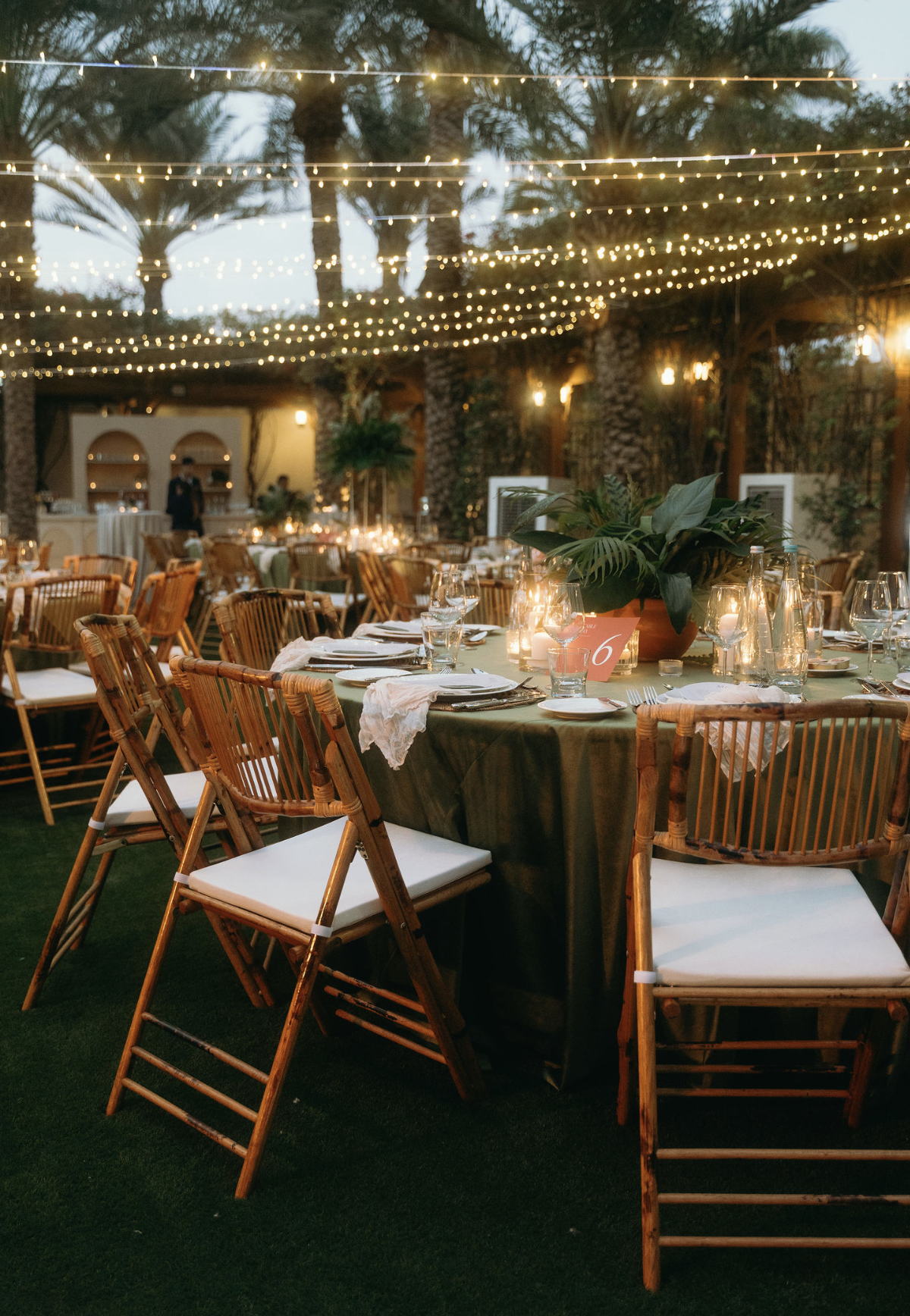A beautifully decorated outdoor event space with bamboo chairs, a round table with green tablecloth, candles, glassware, and a plant centerpiece, illuminated by string lights at dusk.