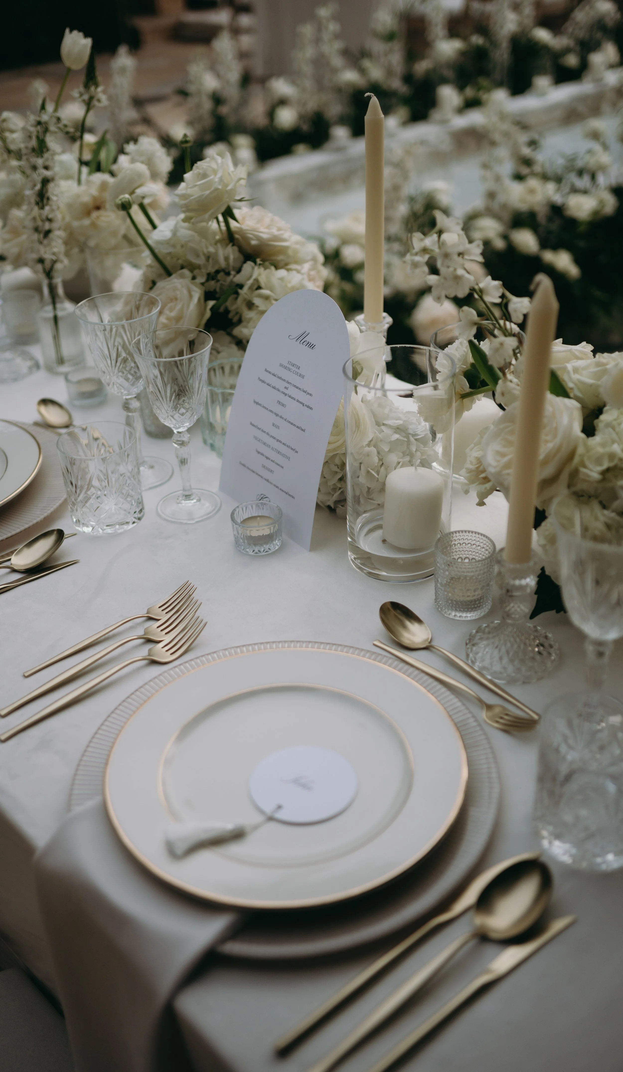 Elegant wedding table setting with gold-rimmed plates, goldcutlery, various empty glasses, white floral centerpieces, tall candles, and a menu card on a white tablecloth.
