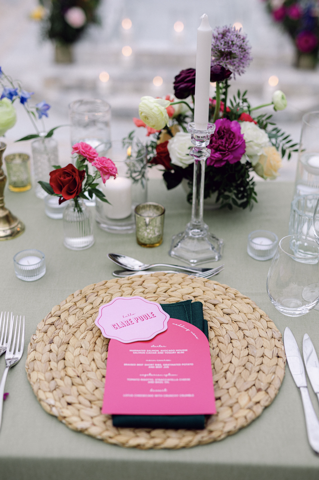 A table setting at an event with a woven placemat, pink menu card, silverware, and glassware. The centerpiece features colorful flowers in a glass vase and a tall white candle on a candlestick. The background includes additional flower arrangements a