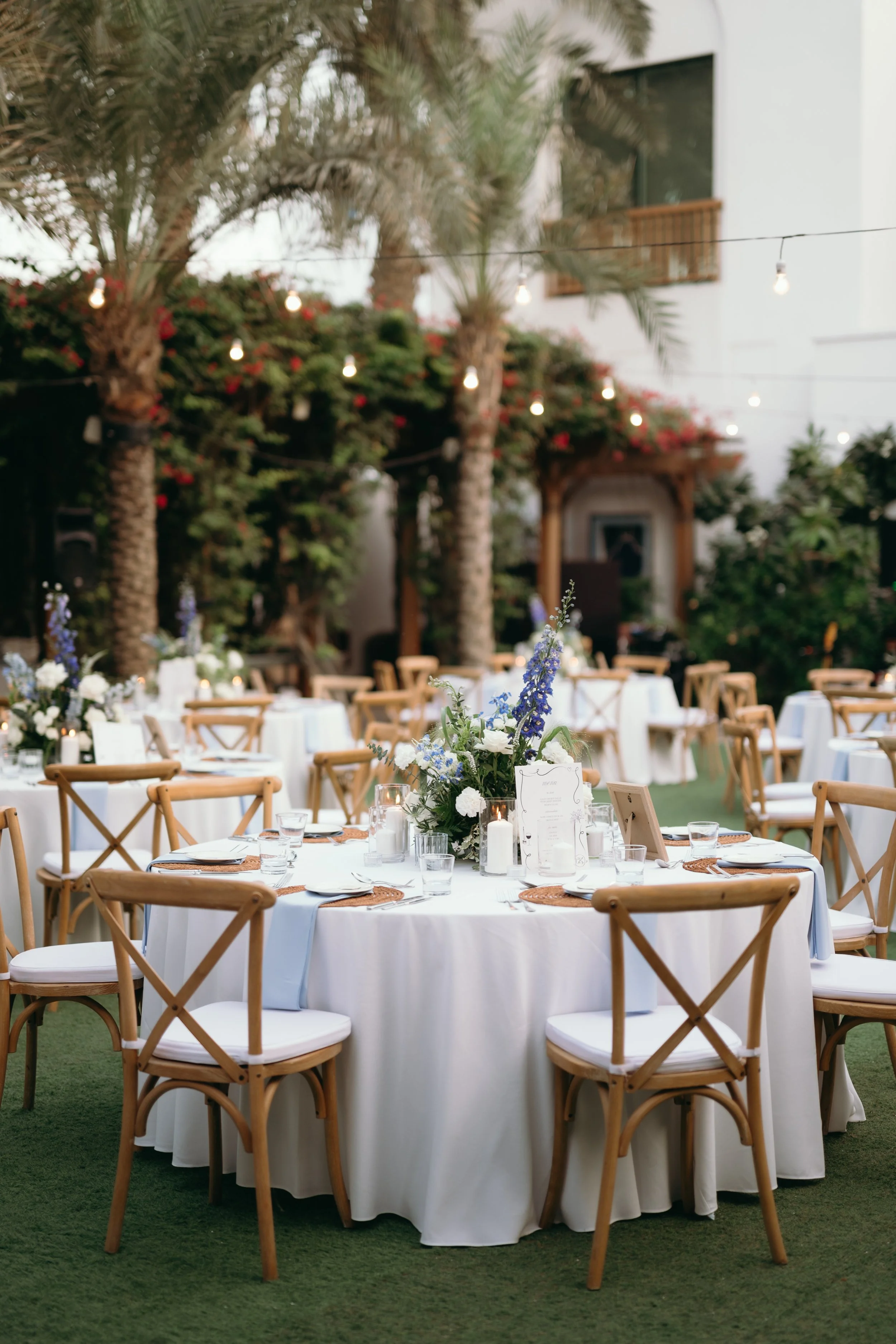 Outdoor wedding reception with round tables covered in white tablecloths, decorated with centerpieces of flowers and candles, surrounded by wooden cross-back chairs, set on green grass, with palm trees and string lights in the background.