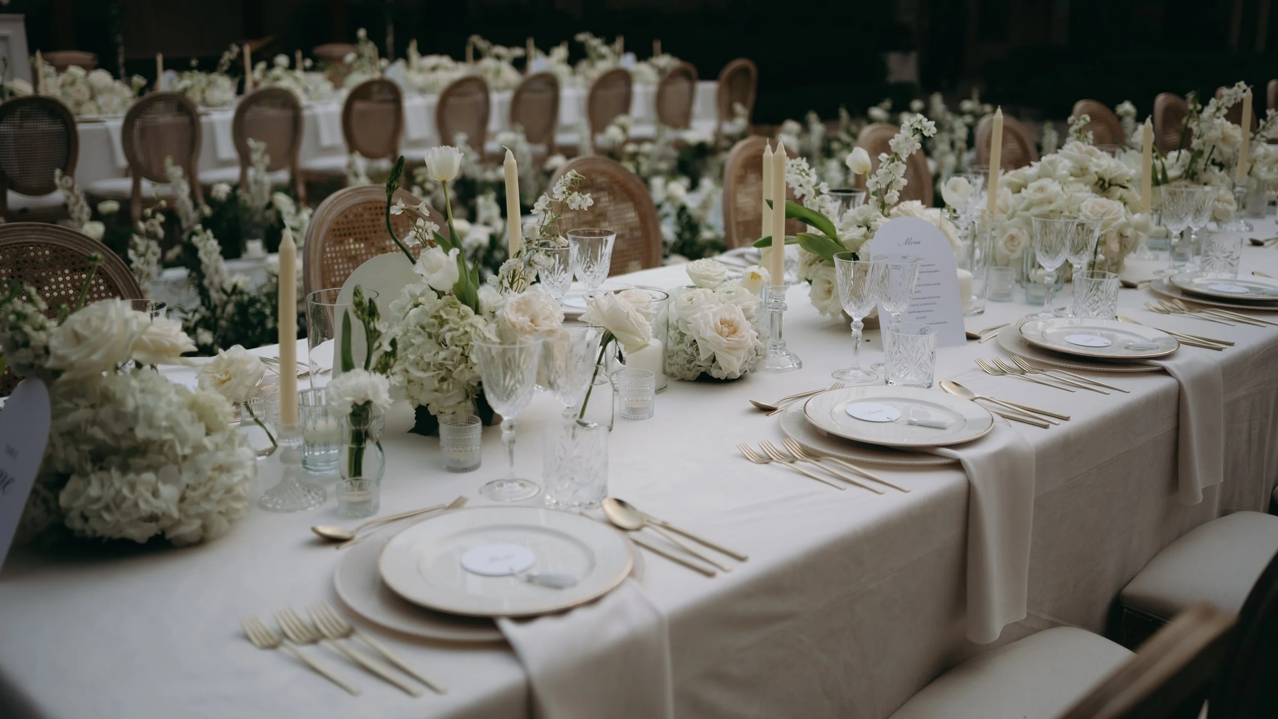 Elegant wedding reception table decorated with white flowers, candles, glassware, and gold cutlery, arranged with place settings and menu cards.