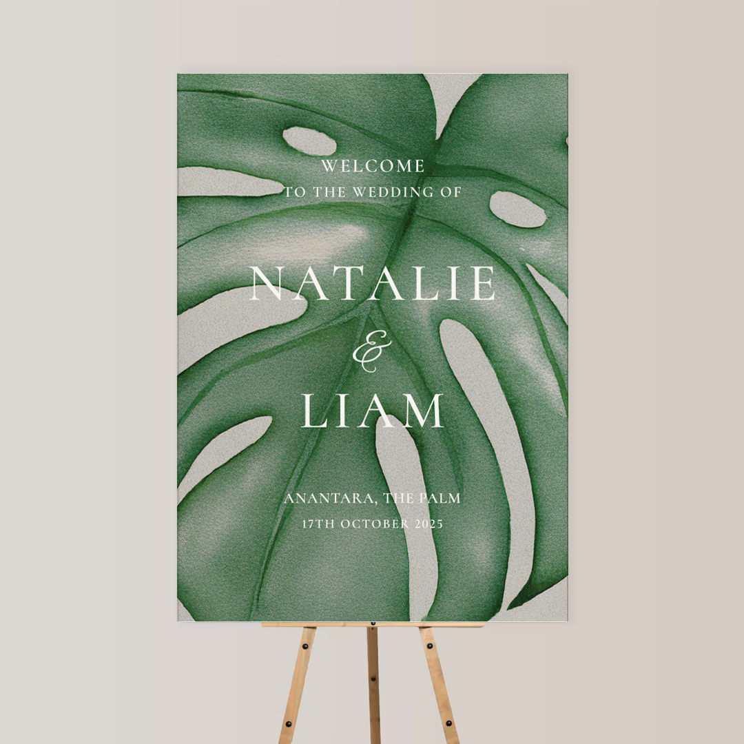 Wedding welcome sign with large tropical green leaves background, displaying text 'Welcome to the wedding of Natalie & Liam, Anantara, The Palm, 17th October 2025' on an easel.