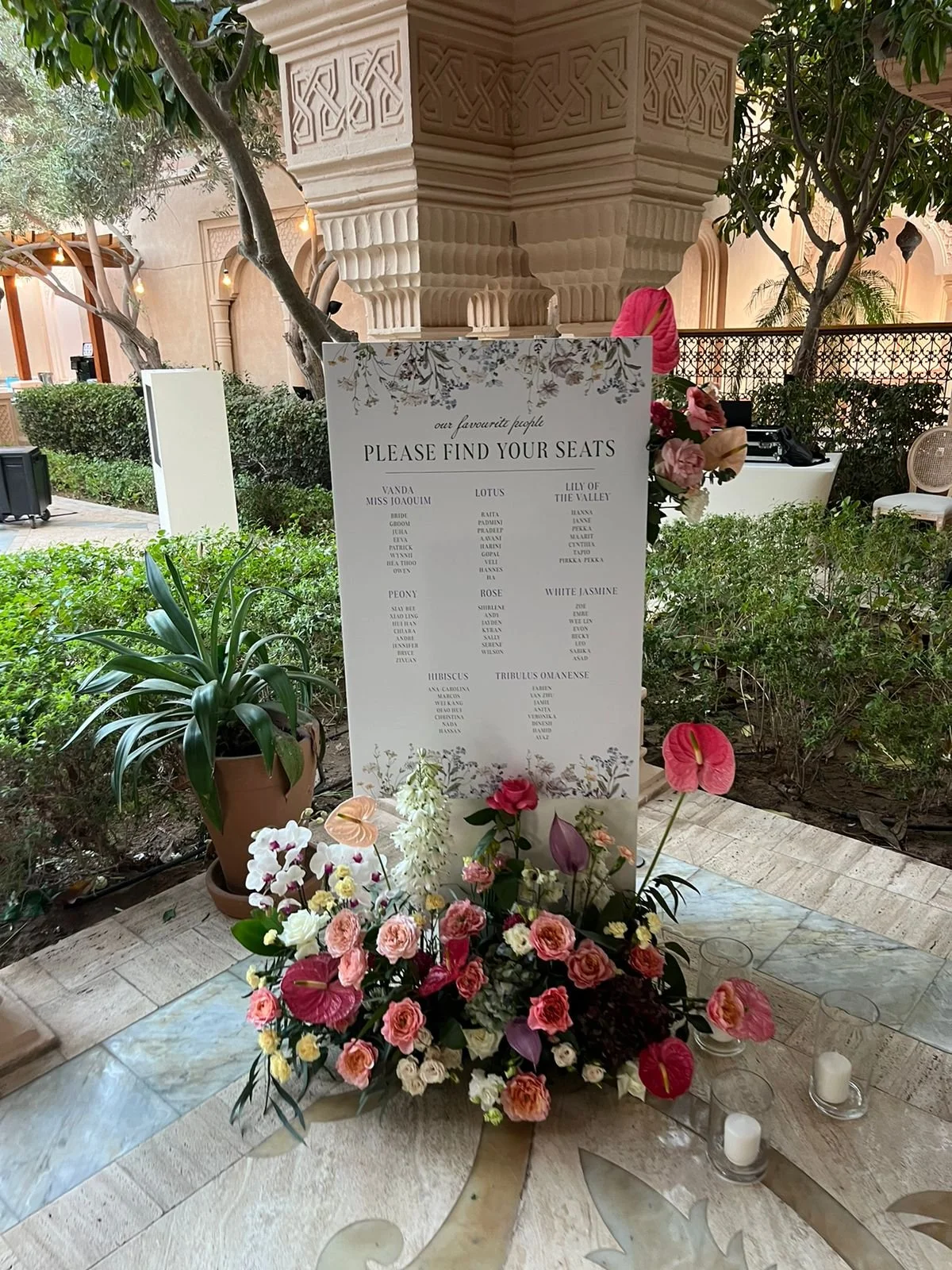 A wedding seating chart displayed on a white board with floral decorations in an outdoor garden setting. The chart lists names under categories such as Vanda, Lotus, Lily of the Valley, Peony, Rose, Hibiscus, and Tribulus Omani, with a floral arrange