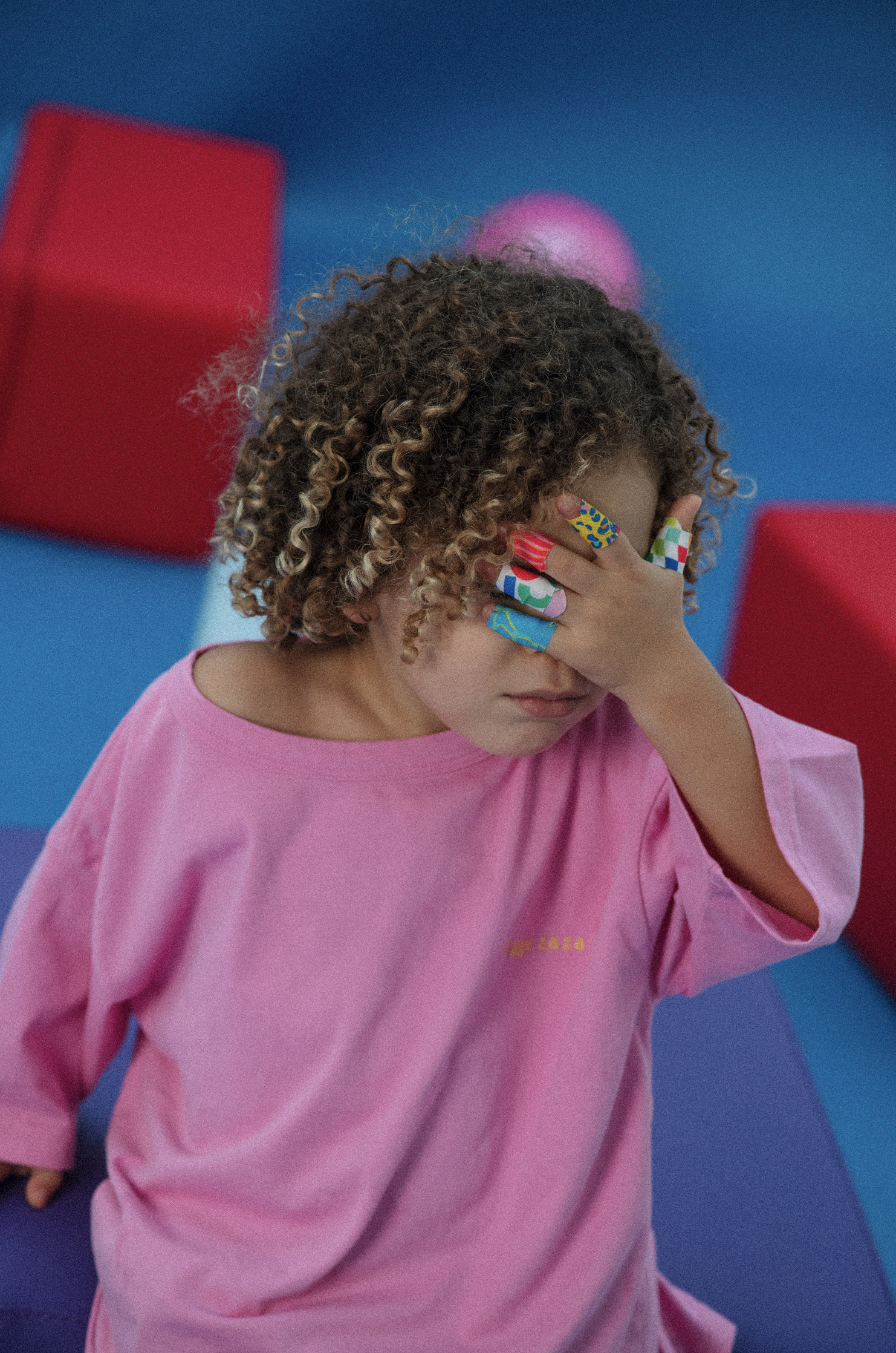 A young girl wearing a pink oversized shirt with colorful tape on her fingers, covering her face with her hand, standing against a blue background with red, purple, and pink geometric shapes.