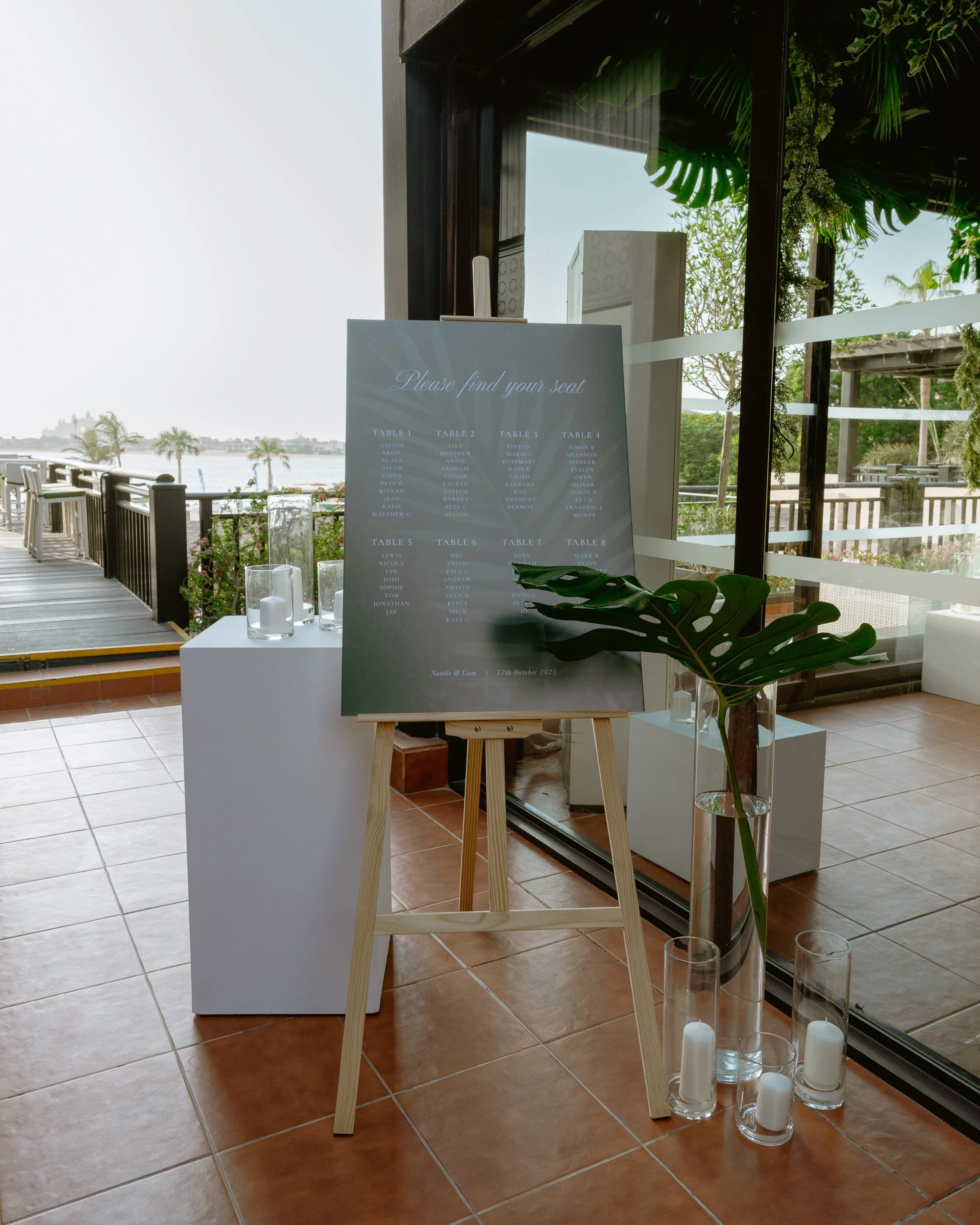 A wedding or event sign listing table assignments with a waterfront view, surrounded by candles and green foliage, in a room with tiled floor and large windows.