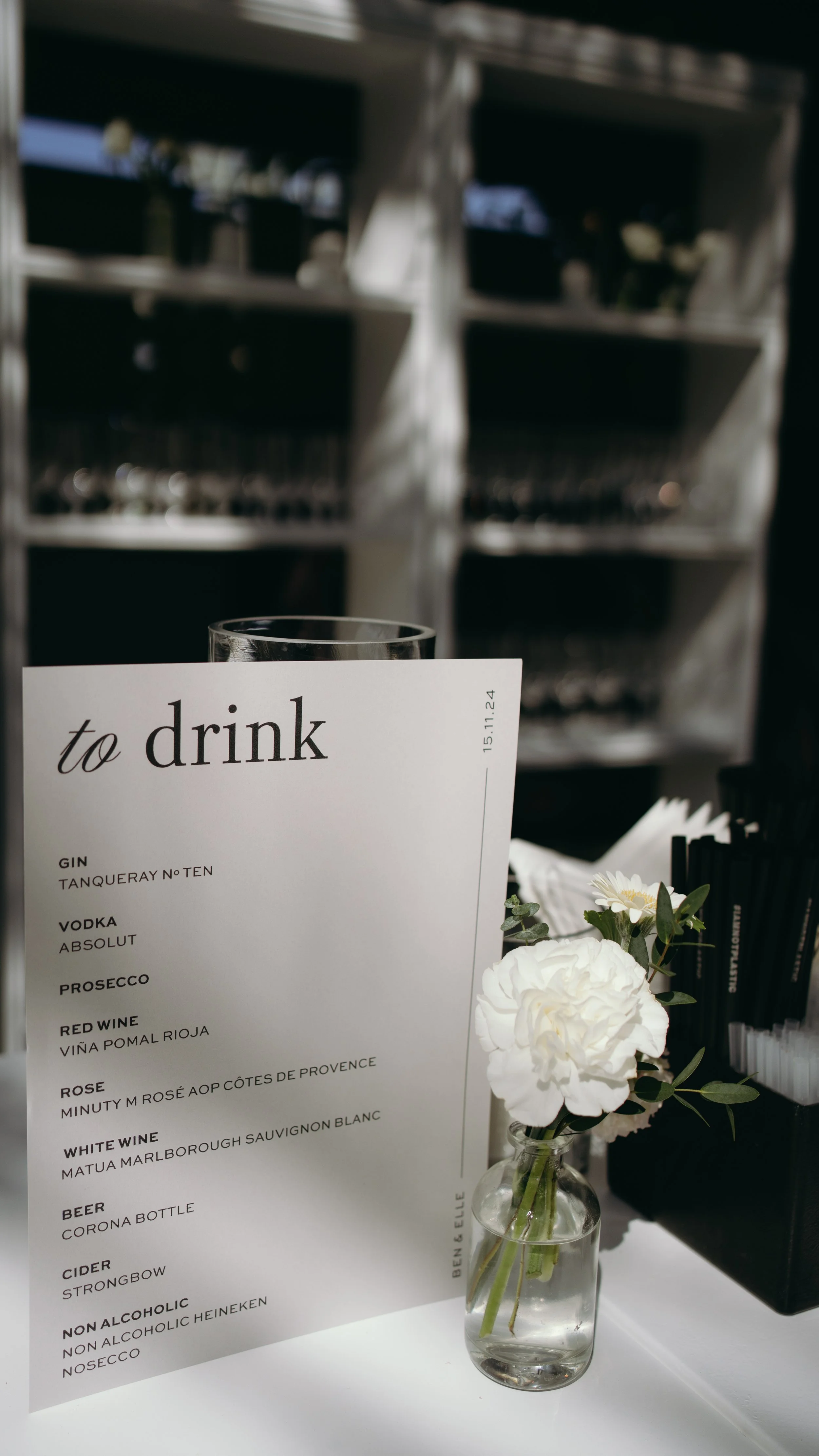 A drink menu on a white sheet of paper and a small bouquet of white flowers in a glass vase on a white table, with a background of metal shelves.
