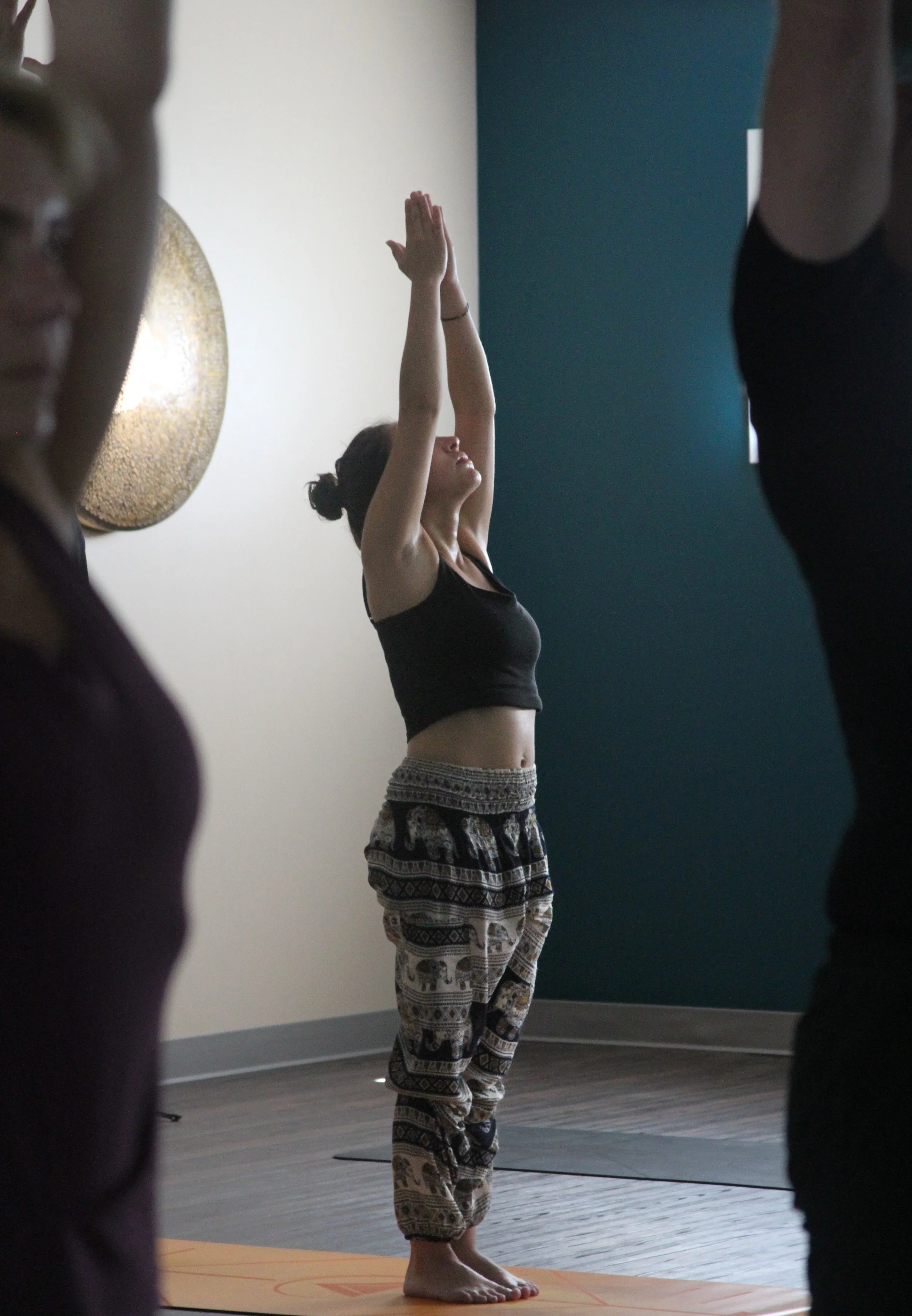 A woman in black yoga pants and a crop top standing on a yoga mat with her eyes closed and hands raised above her head in a yoga pose, surrounded by other people in a yoga class.