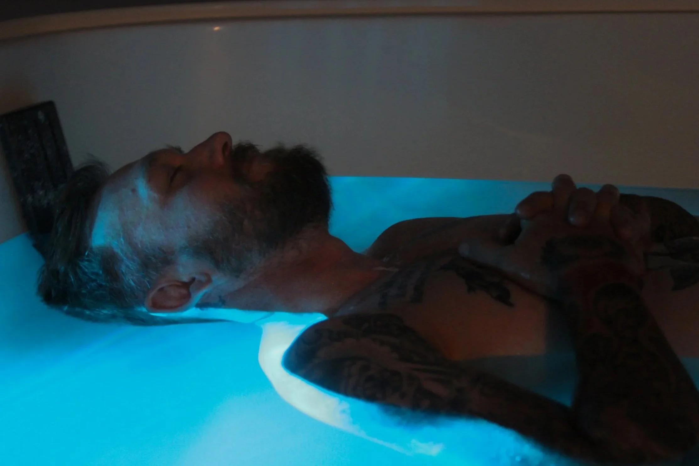 A person with a beard and tattoos lying down with closed eyes, resting their head on a white pillow, with their hand resting on their chest, under a soft blue light.