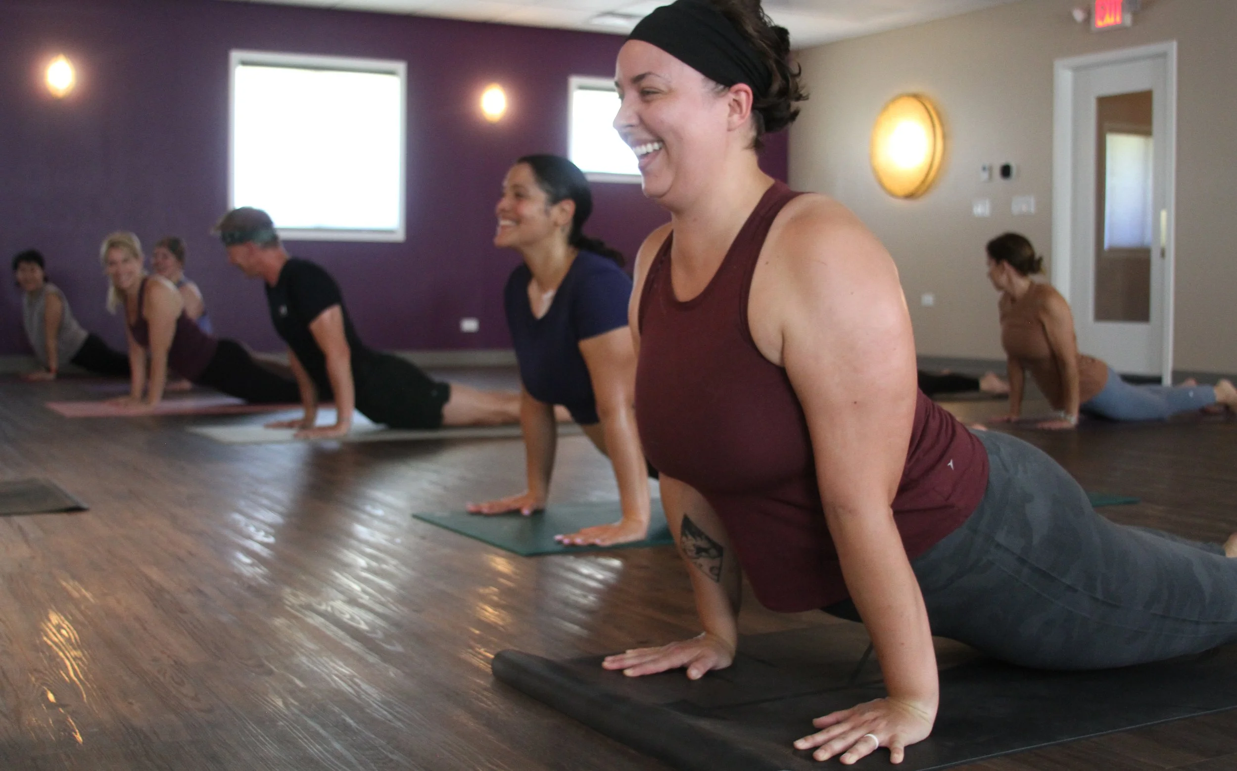 Group of women participating in a yoga class on mats, in a room with purple and beige walls, smiling and stretching in a yoga pose.