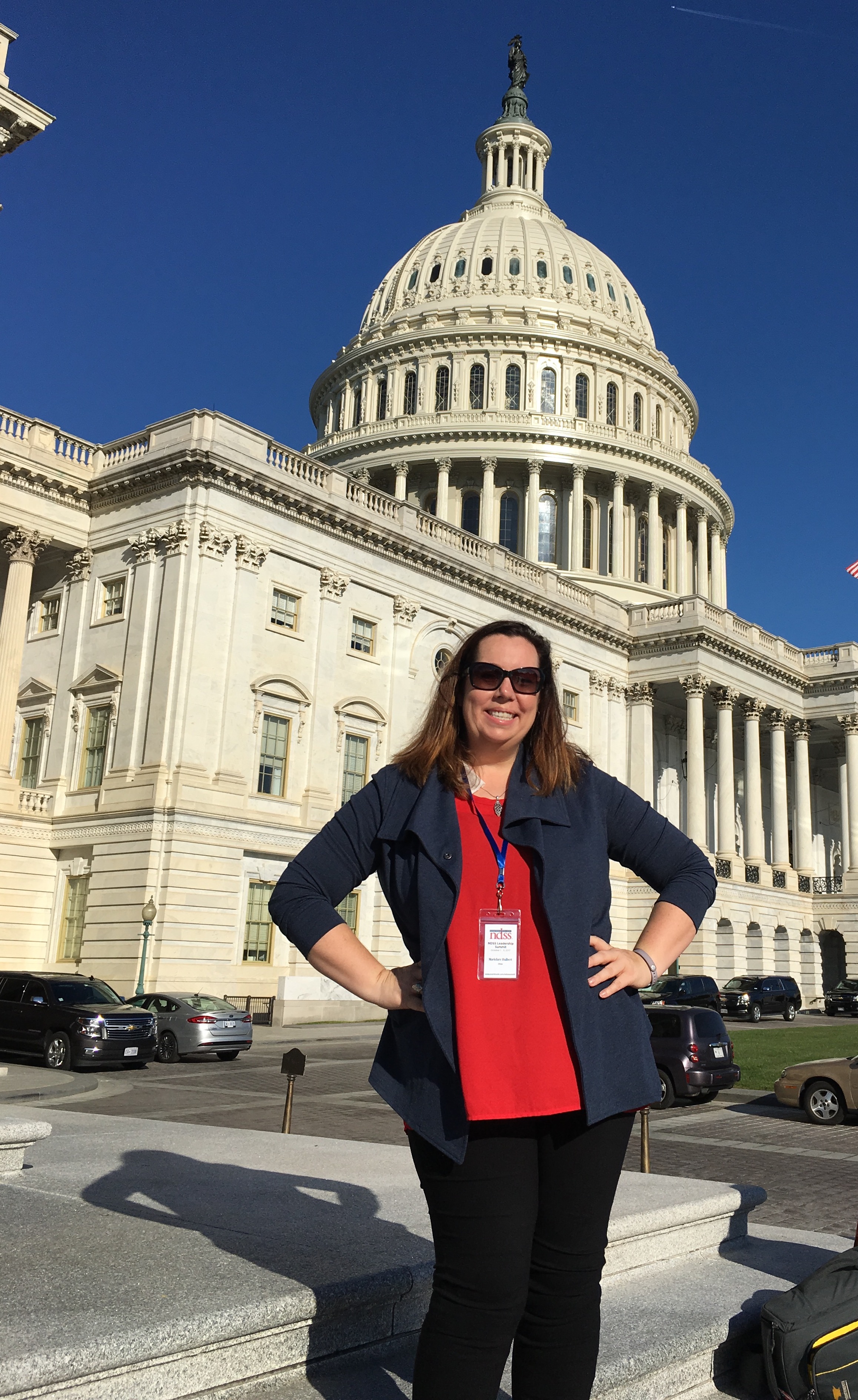 Alt Text: A white woman stands proudly in front of a large white ornate building on Capitol Hill. She wears a red shirt,  blue blazer and sunglasses. She smiles. A visitor pass hangs at her neck