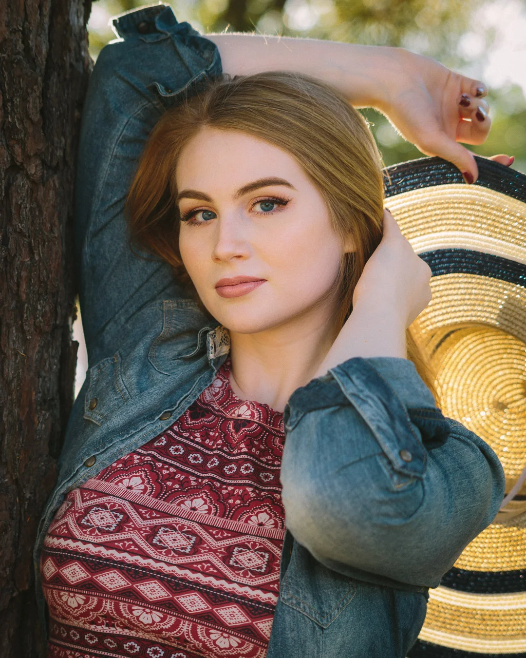 senior leaning on tree with denim jacket and hat