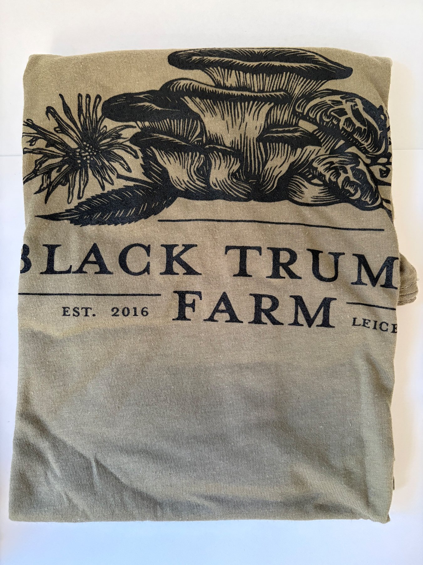 This folded cotton t-shirt depicts a bouquet of mycelium, such as oyster and shitake mushrooms, printed in black ink on green fabric.