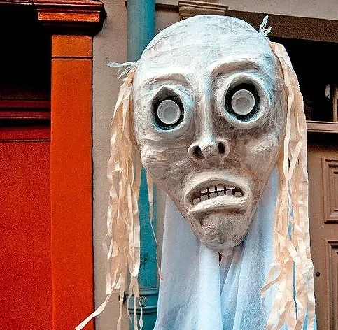 A Halloween decoration featuring a ghostly face with large, round, hollow eyes and a distressed expression, with long, tattered paper-like hair.