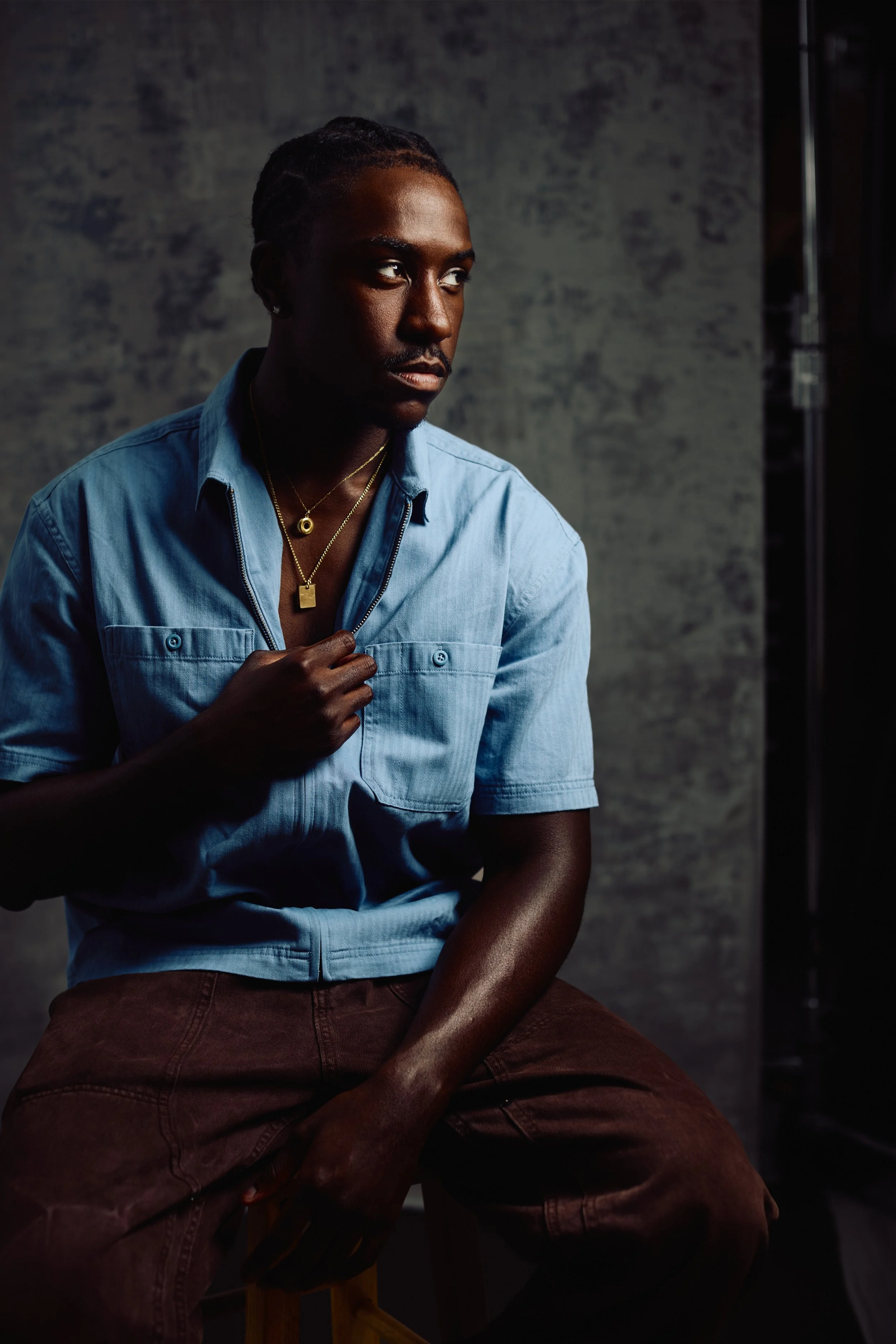 A man with dark skin and braided hair, wearing gold necklaces and a light blue shirt, is looking to his left against a dark, textured background.