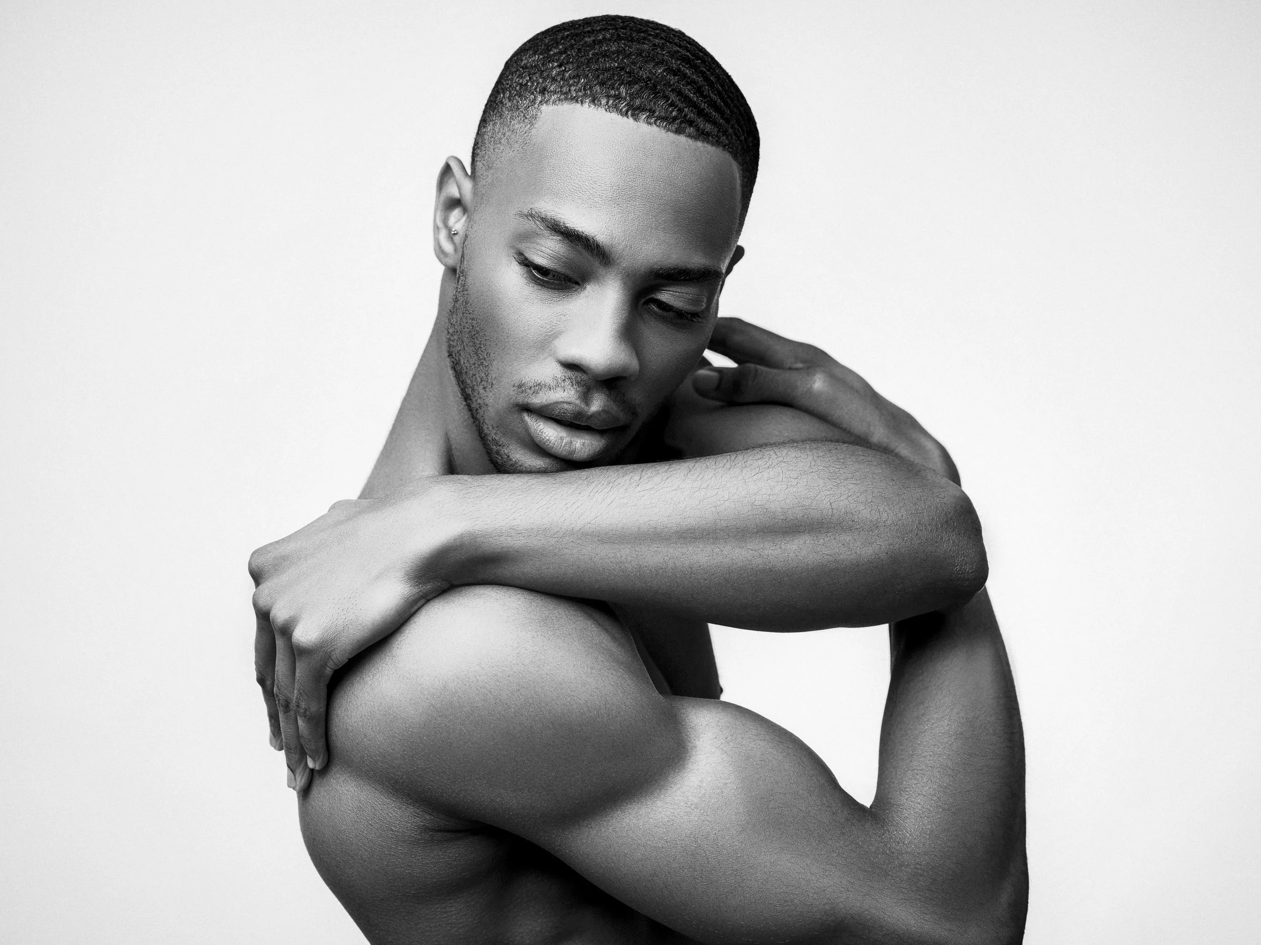 Black and white portrait of a young man with short hair, looking downward, with arms crossed over his shoulders, arms and head resting on them.