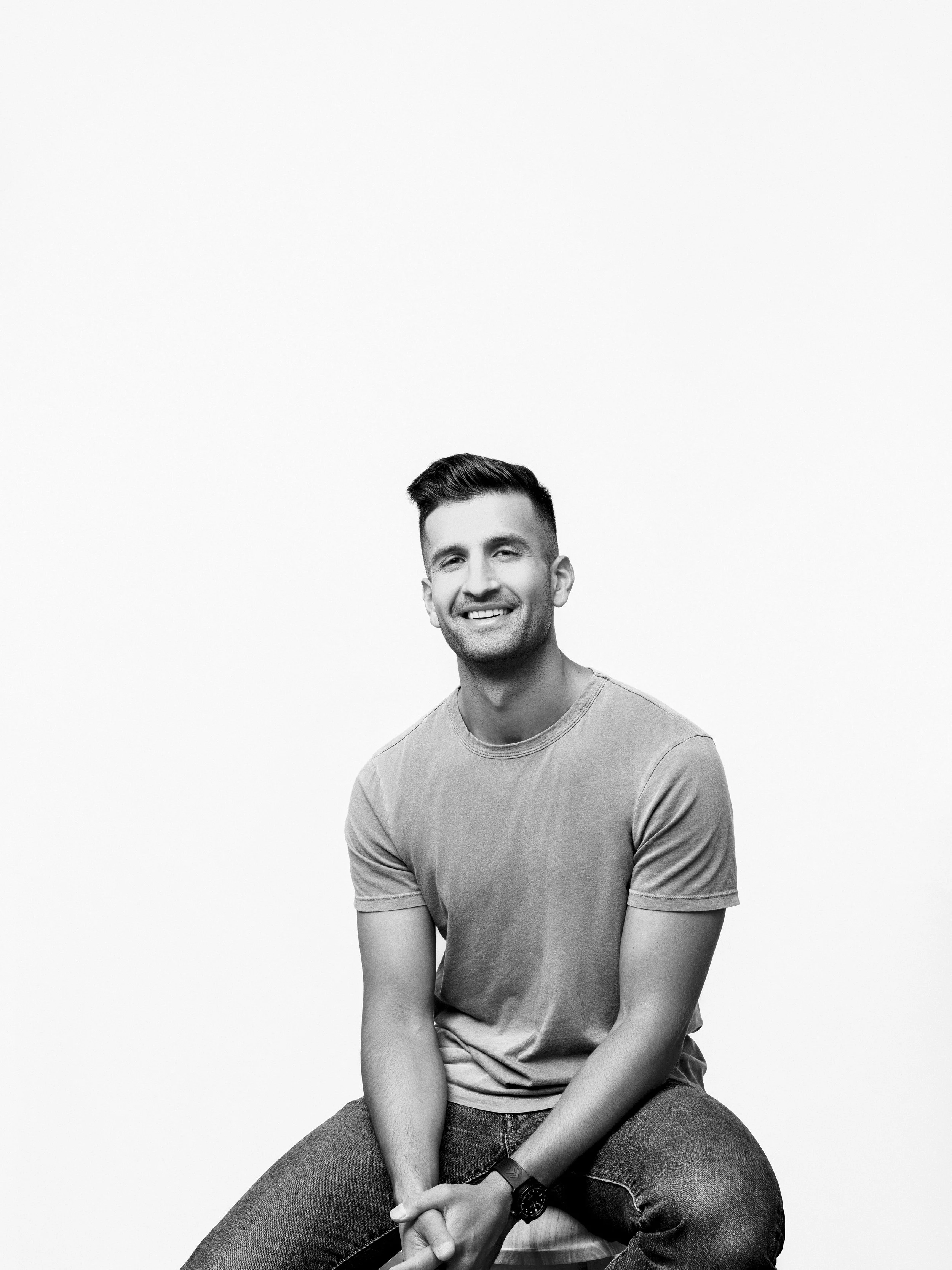 A young man with dark hair and a beard, smiling, sitting on a stool in casual clothing, in front of a plain white background.