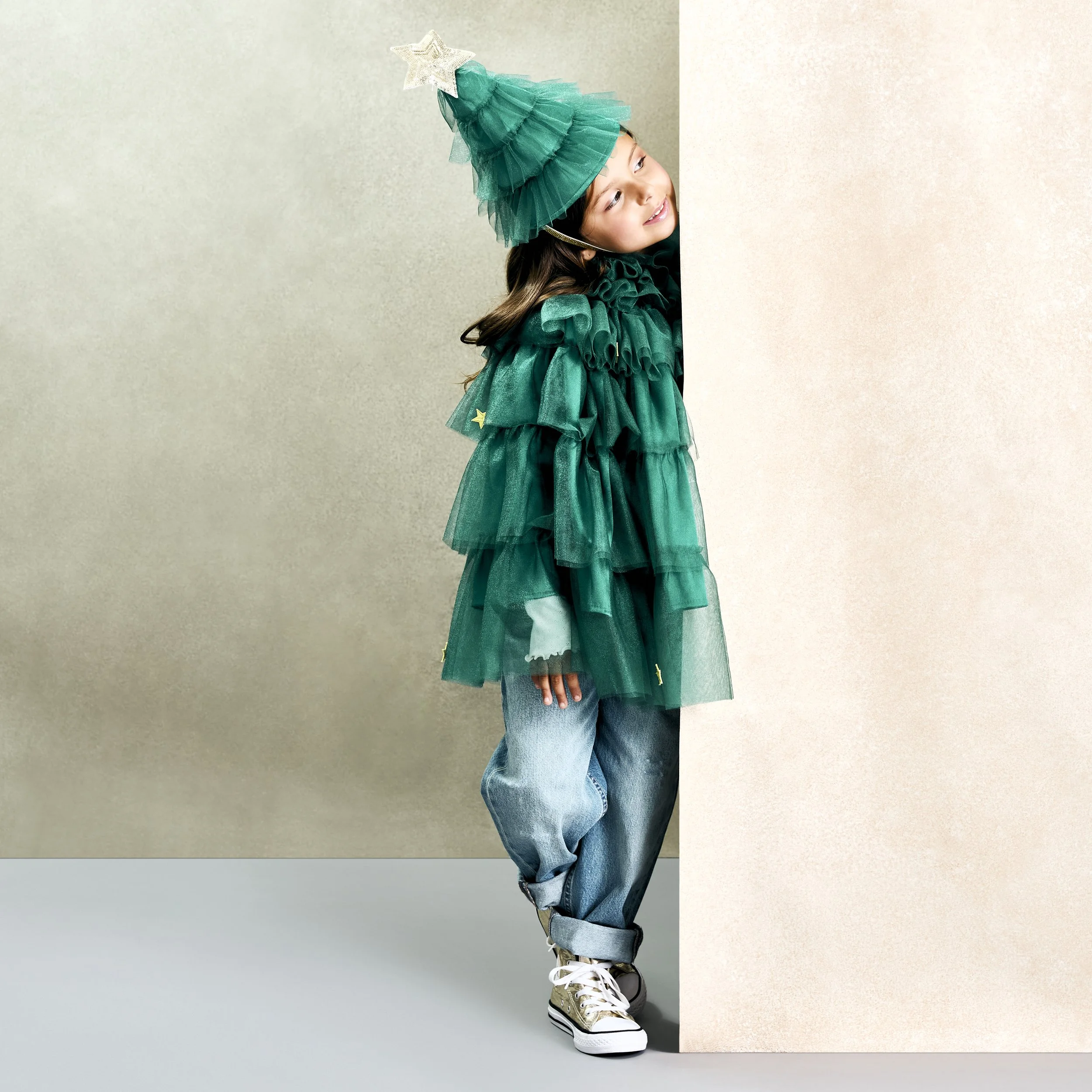 Young girl dressed as a Christmas tree peeking out from behind a beige wall, wearing a green tiered tutu dress and a matching green Christmas tree hat with a silver star on top, and white sneakers.