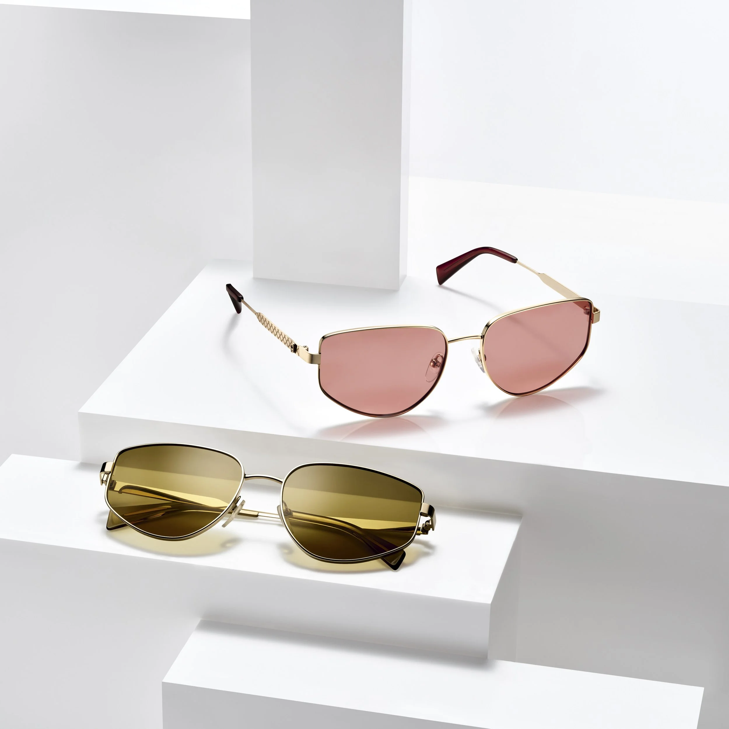 Two pairs of sunglasses, one with pink lenses and gold frames, and the other with yellow lenses and gold frames, displayed on white geometric platforms.