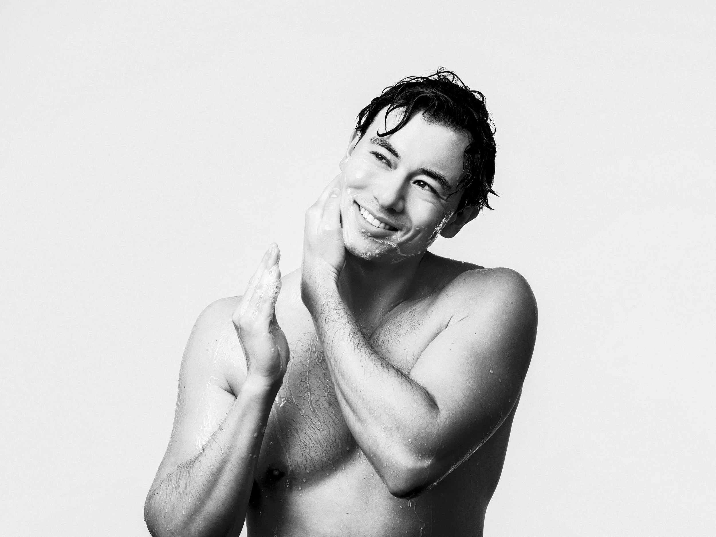 A young man with wet hair is smiling and washing his face with soap in a black and white photo.