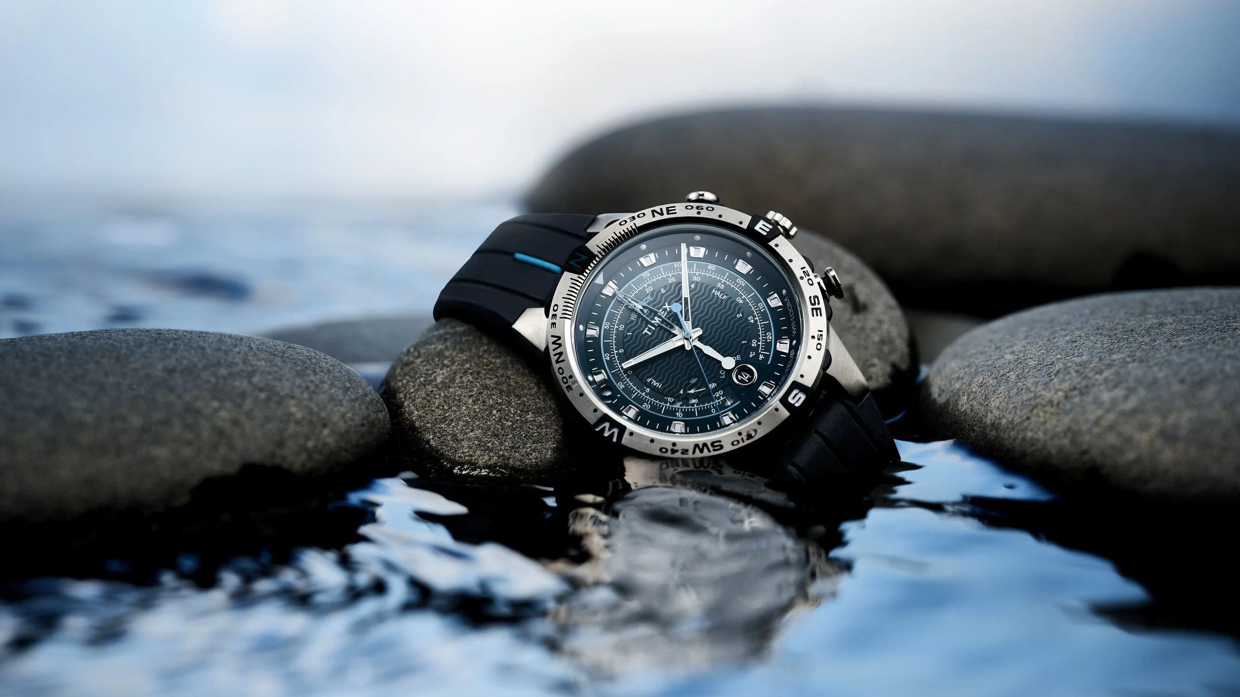 A black and silver wristwatch with a detailed blue dial resting on a pile of smooth stones near water.