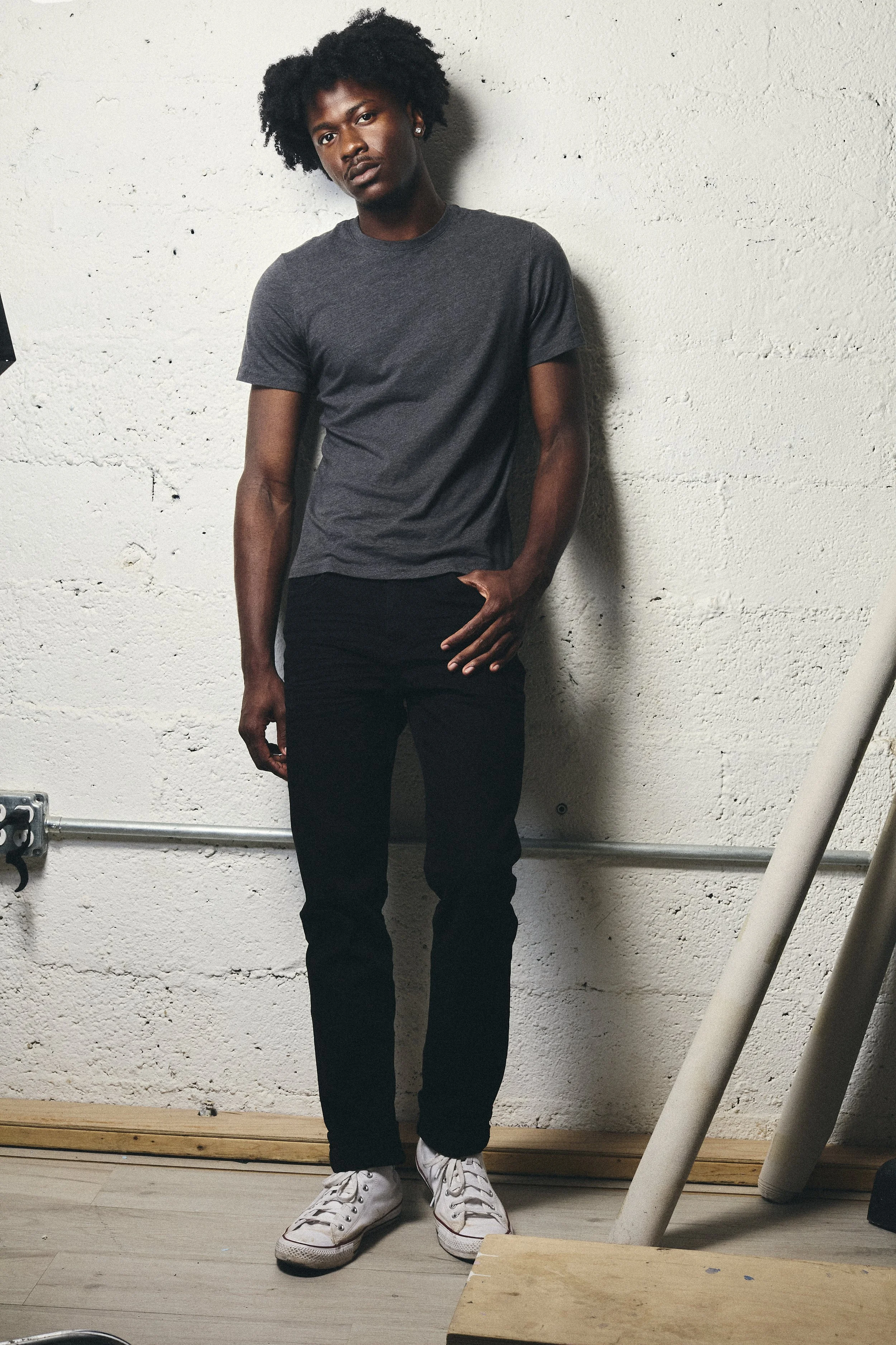 Young man with curly black hair wearing a gray T-shirt, black pants, and white sneakers standing against a textured white wall.
