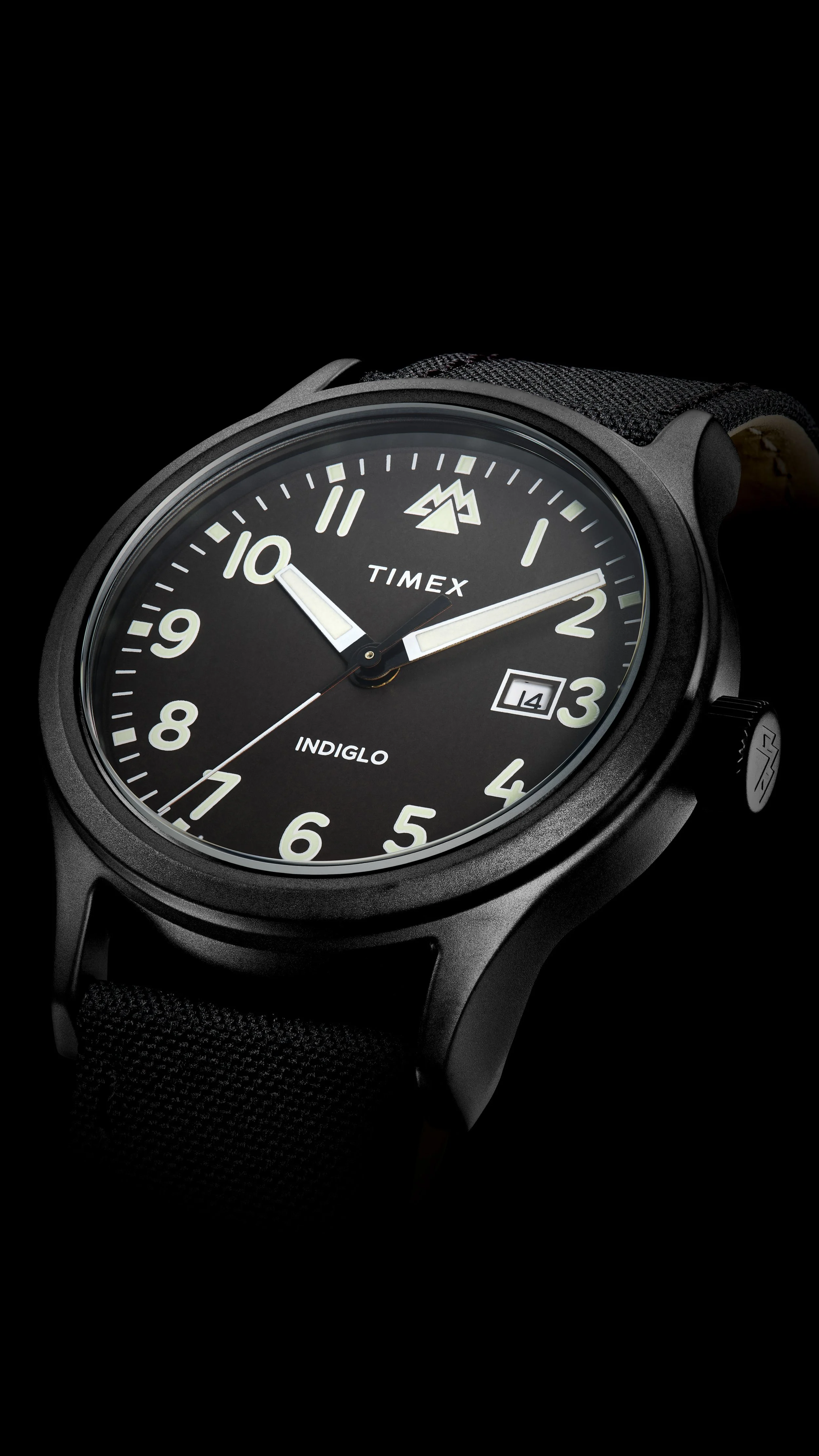 Close-up of a black Timex watch with a black dial, Arabic numerals, luminous hands, a date display at 3 o'clock, and the INDIGLO feature, against a dark background.