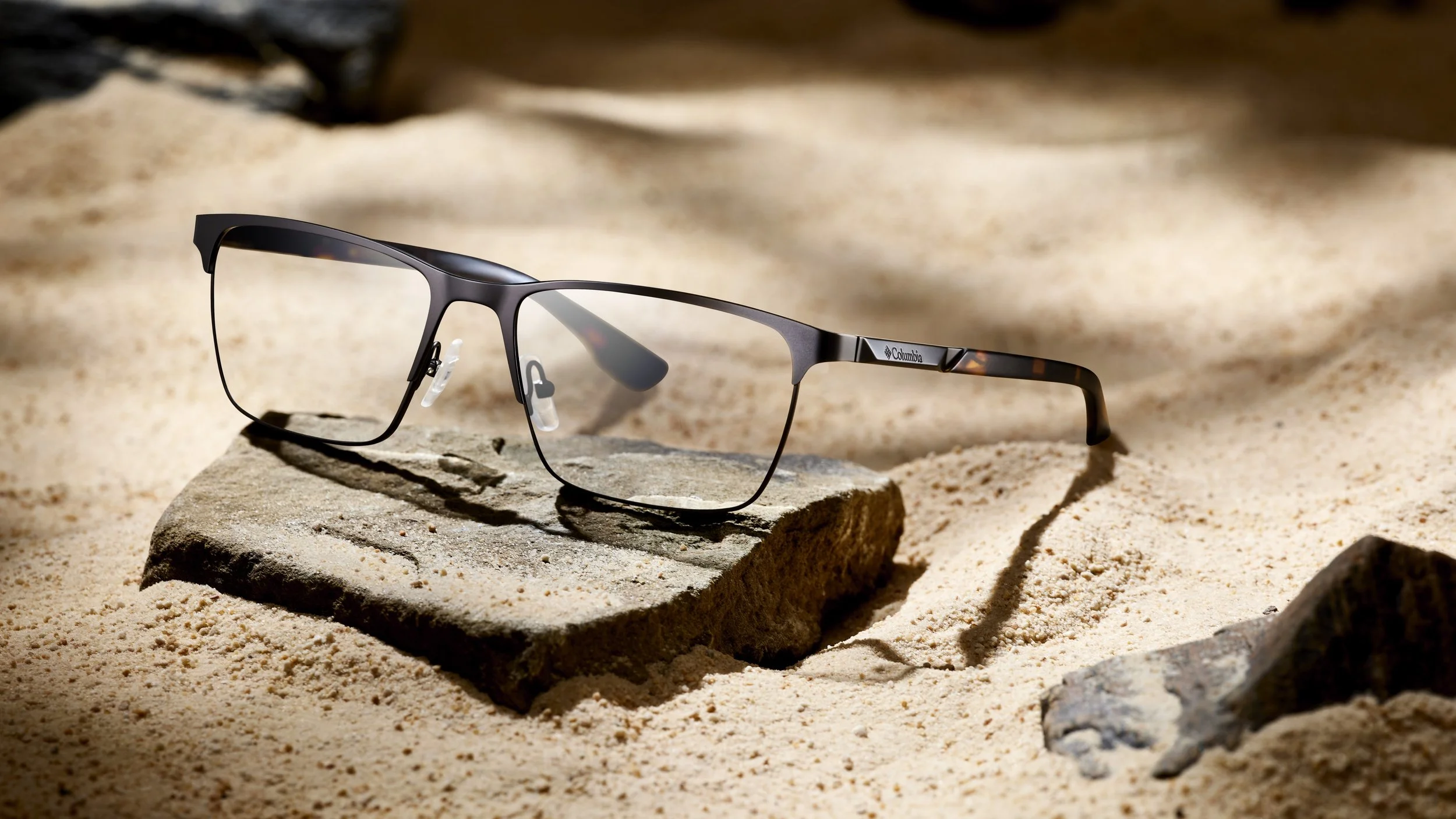 A pair of black Columbia eyeglasses resting on a rock amidst sand and small rocks.