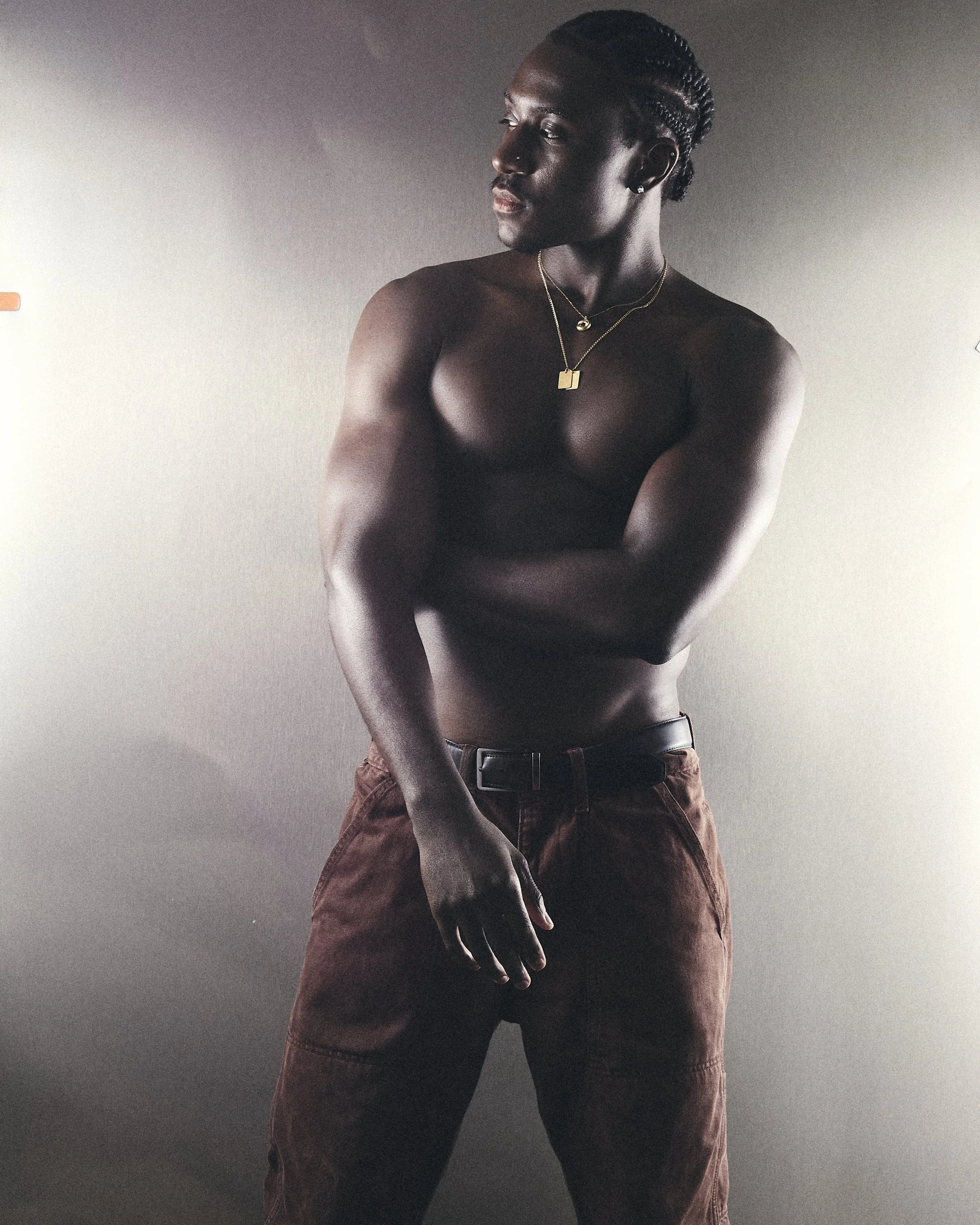 A person with dark skin and short braids stands against a plain background, topless with only brown pants and accessories, looking to the side with arms crossed.