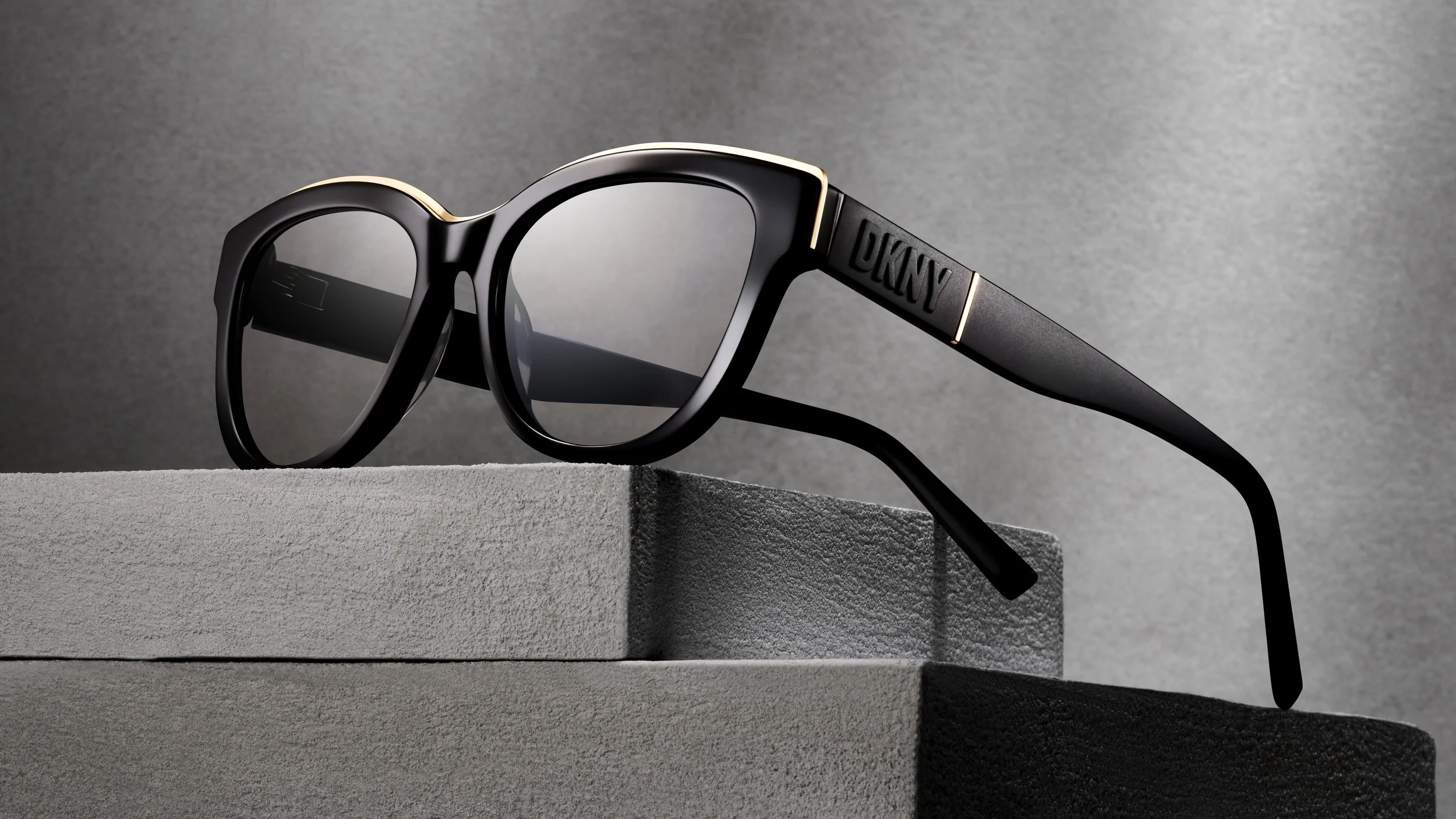 Black sunglasses with gold accents on the frame resting on gray stacked boxes, with a gray textured background.