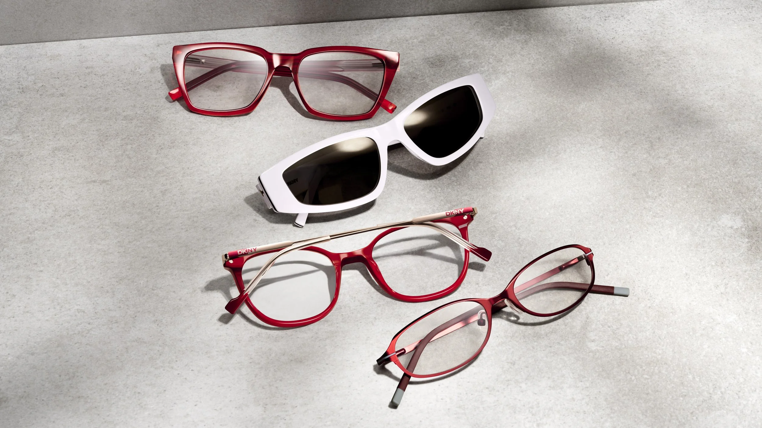 Four pairs of sunglasses, with three in red and one in white, are arranged on a textured, light gray surface, some overlapping each other.