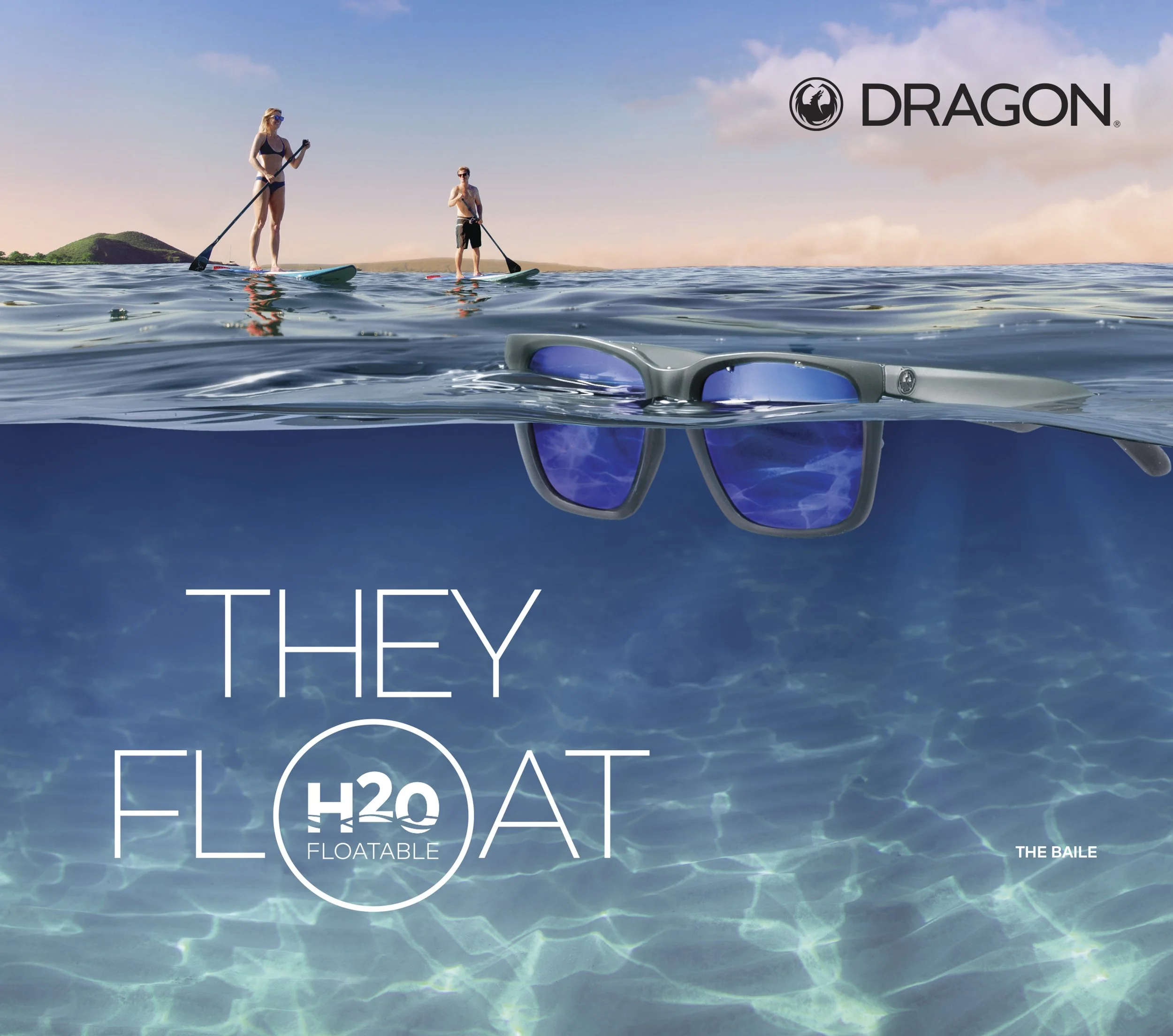 A waterproof pair of sunglasses floating on the water surface of a lake with people paddleboarding in the background and a distant landmass under a blue sky.