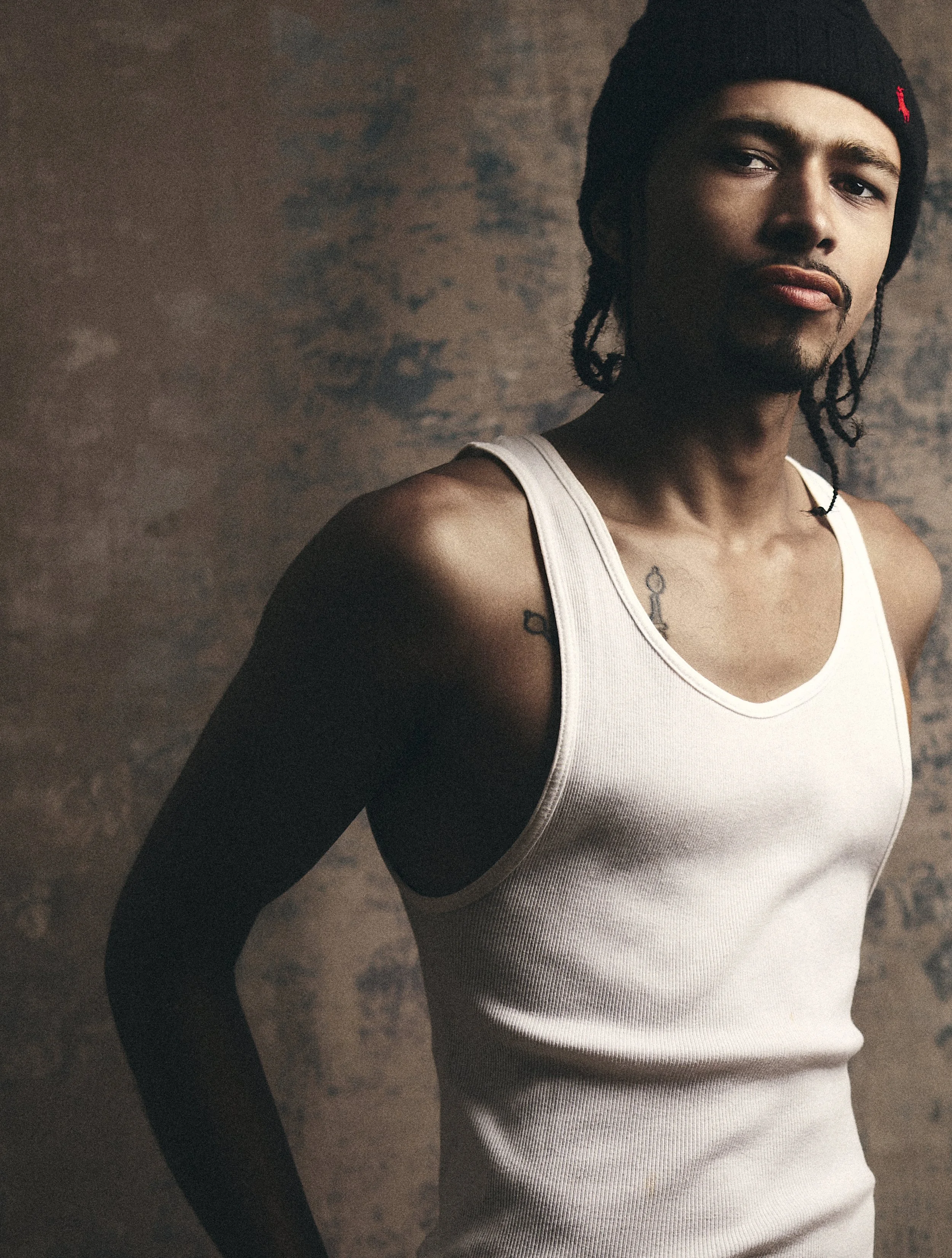 A young man with long braided hair, wearing a black beanie and a sleeveless white shirt, standing against a brick wall.