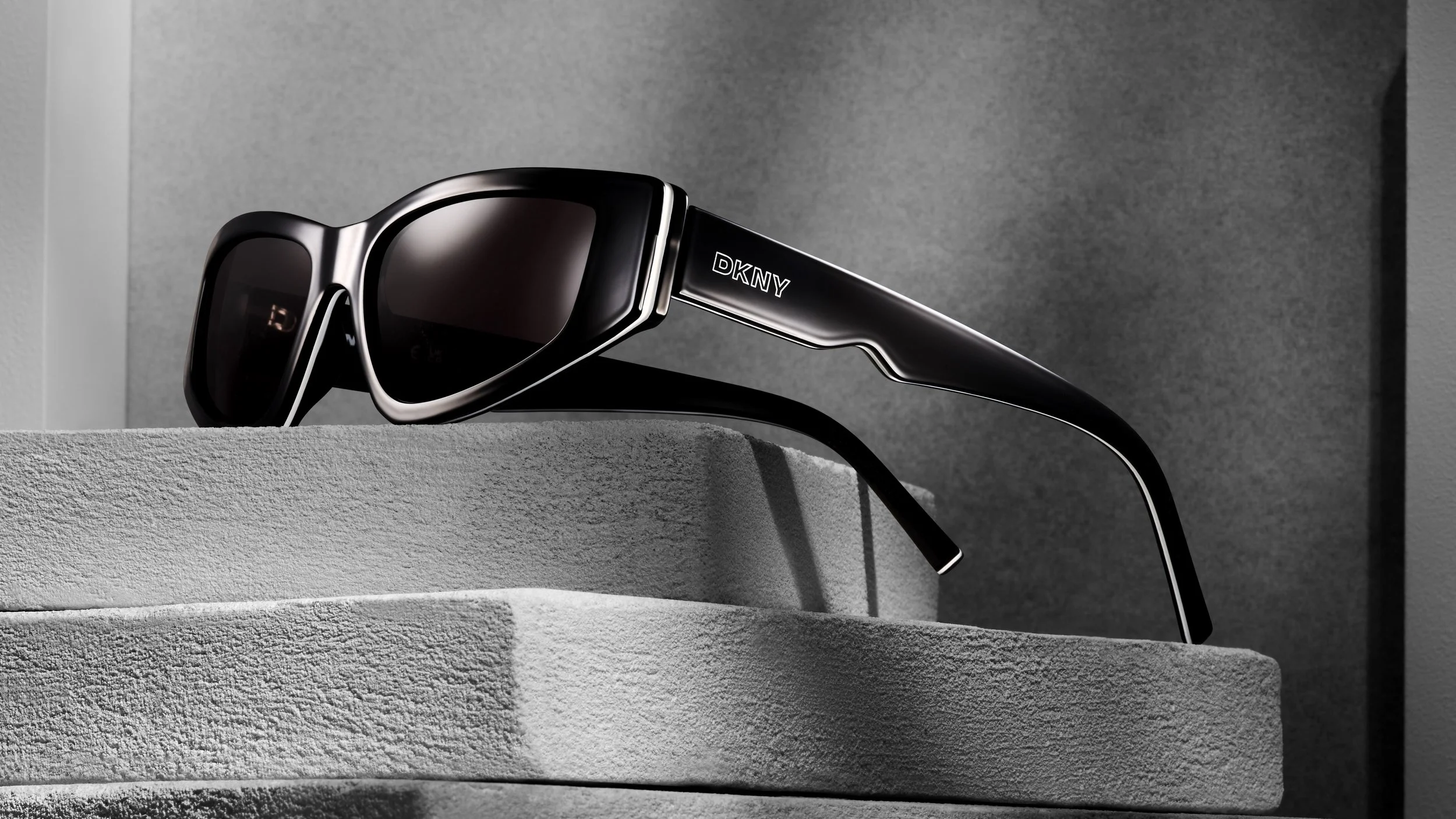 Black DKNY sunglasses with dark lenses resting on textured concrete surfaces against a gray background.