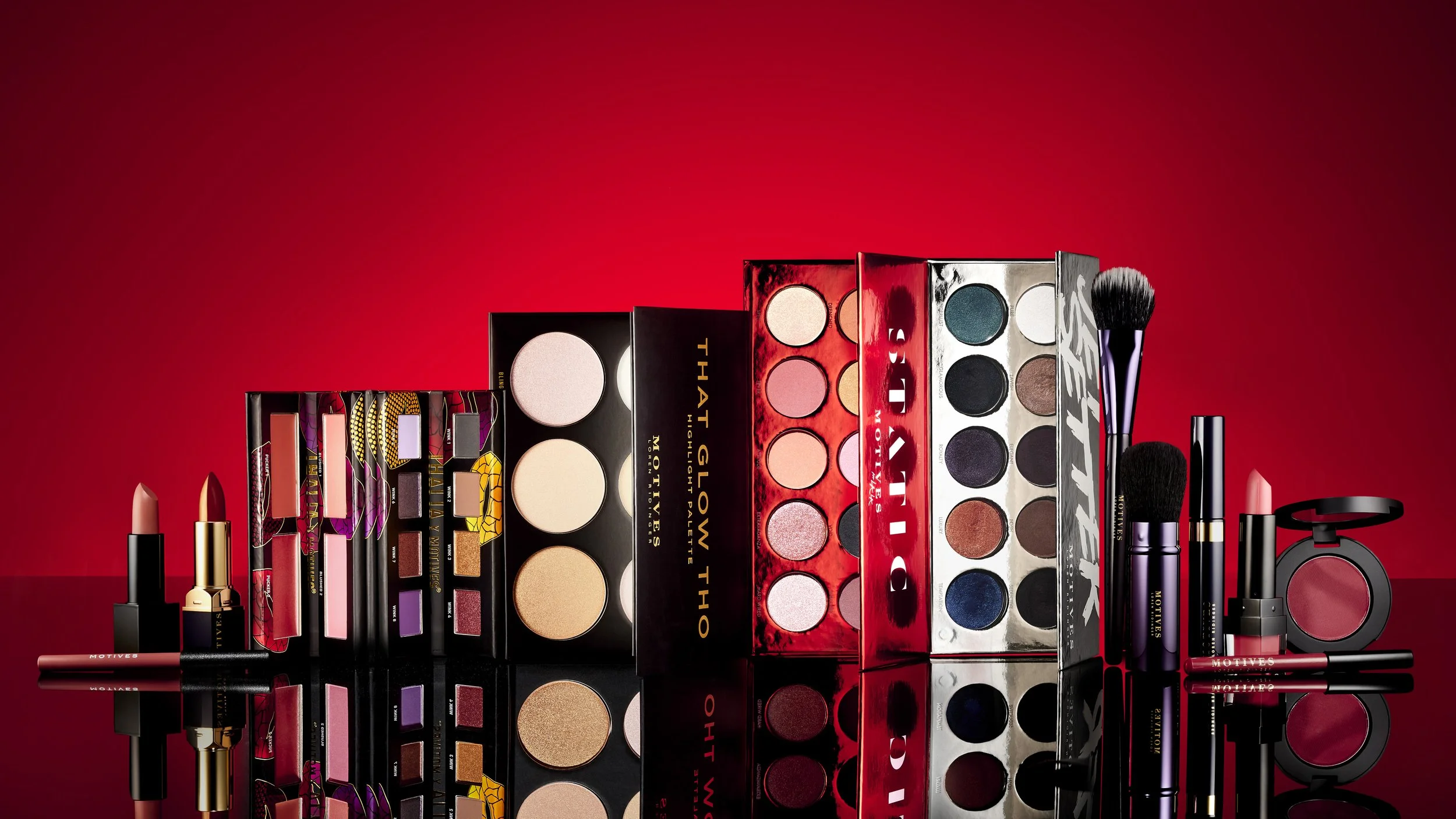 Collection of makeup products including lipsticks, eyeshadow palettes, blush, and brushes arranged in front of a red background.