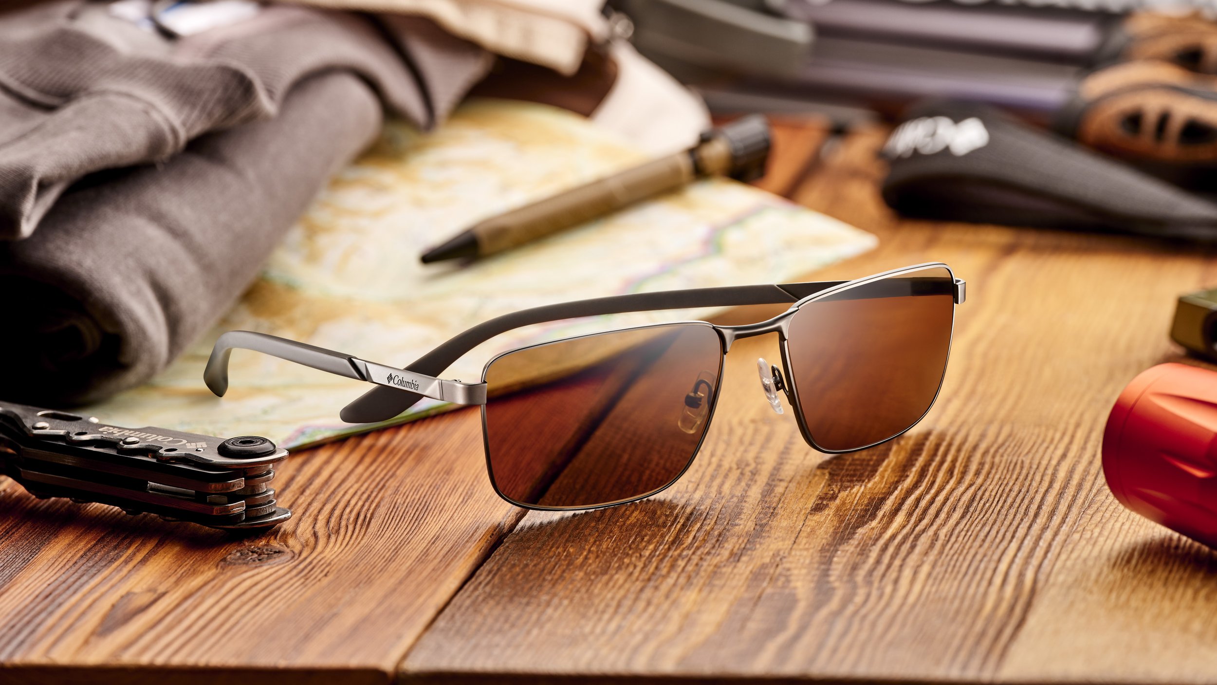 A pair of sunglasses with brown tinted lenses on a wooden table surrounded by outdoor gear including a multi-tool, a map, a pen, a rolled-up shirt, and a wallet.