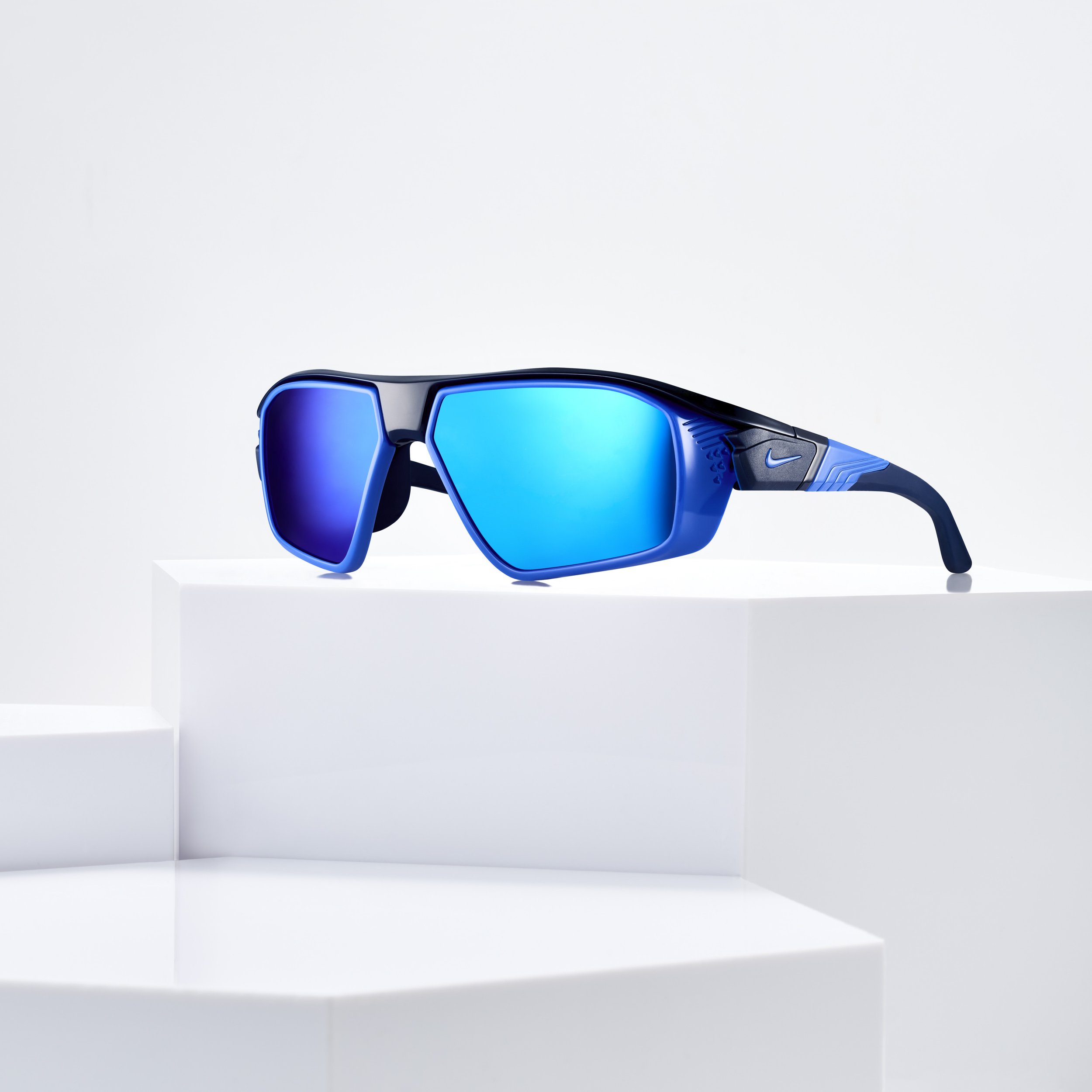 Pair of black and blue Nike sunglasses with mirrored blue lenses displayed on white geometric platforms.