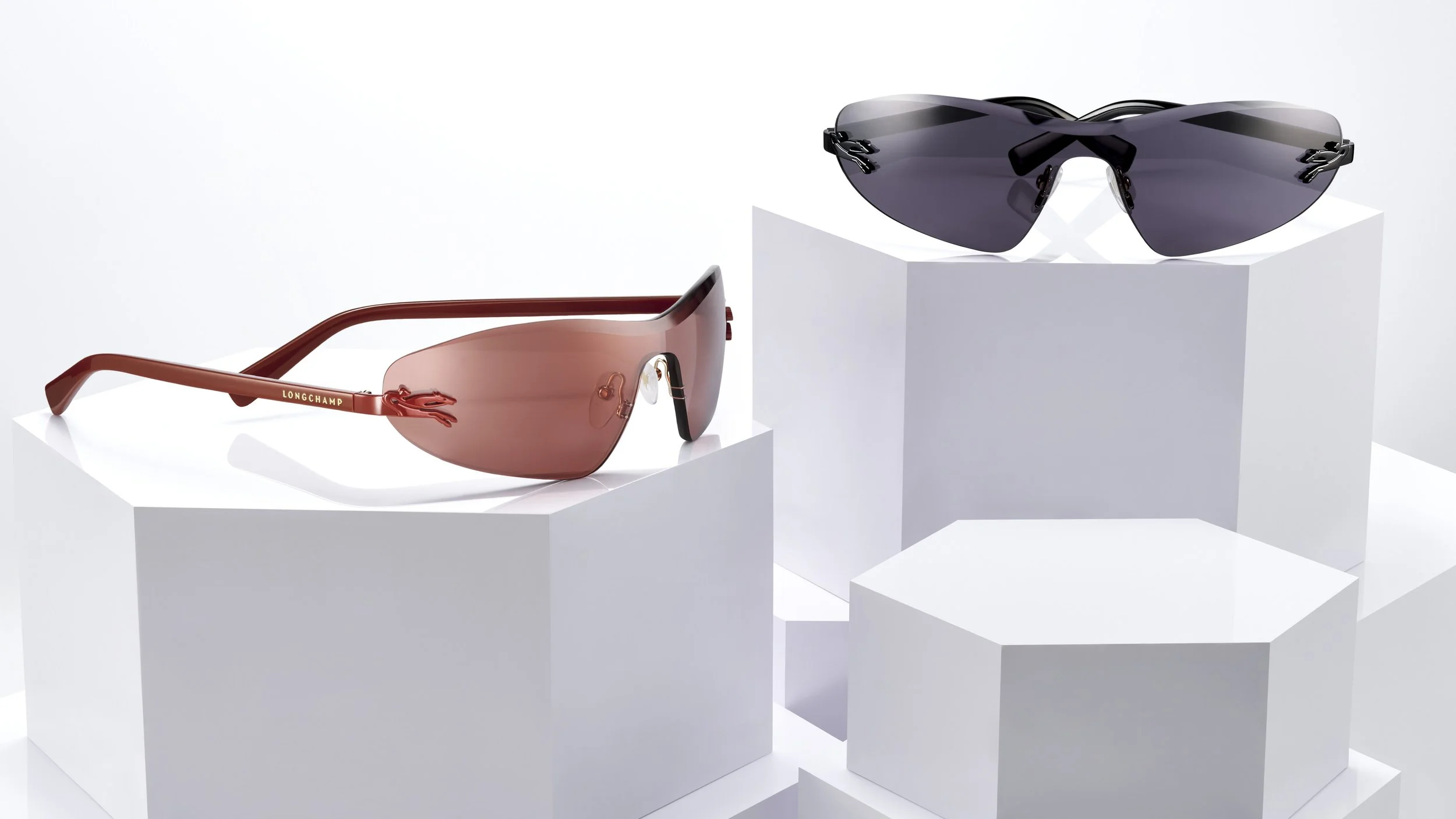 Two pairs of designer sunglasses, one with brown lenses and red frames, the other with dark lenses and black frames, displayed on white platform blocks