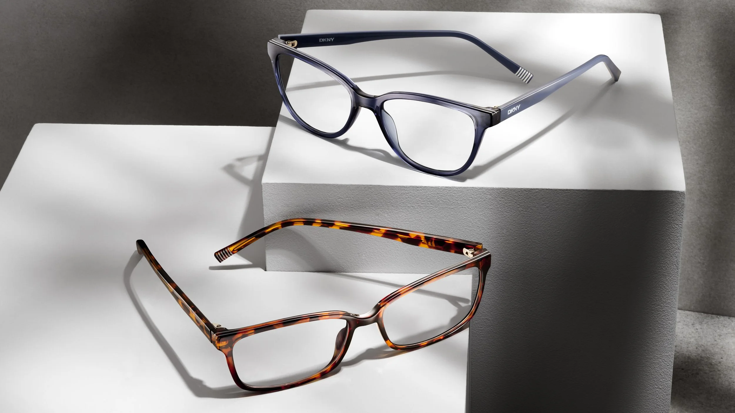 Two pairs of eyeglasses, one with purple frames and the other with tortoiseshell frames, positioned on white and gray textured platforms.