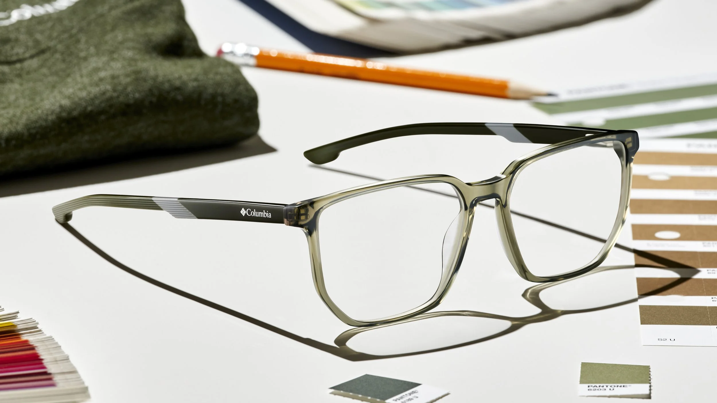 A pair of Columbia eyeglasses on a white surface with color swatches, a color sample chip, a pencil, a green cloth, and fabric swatches in the background.