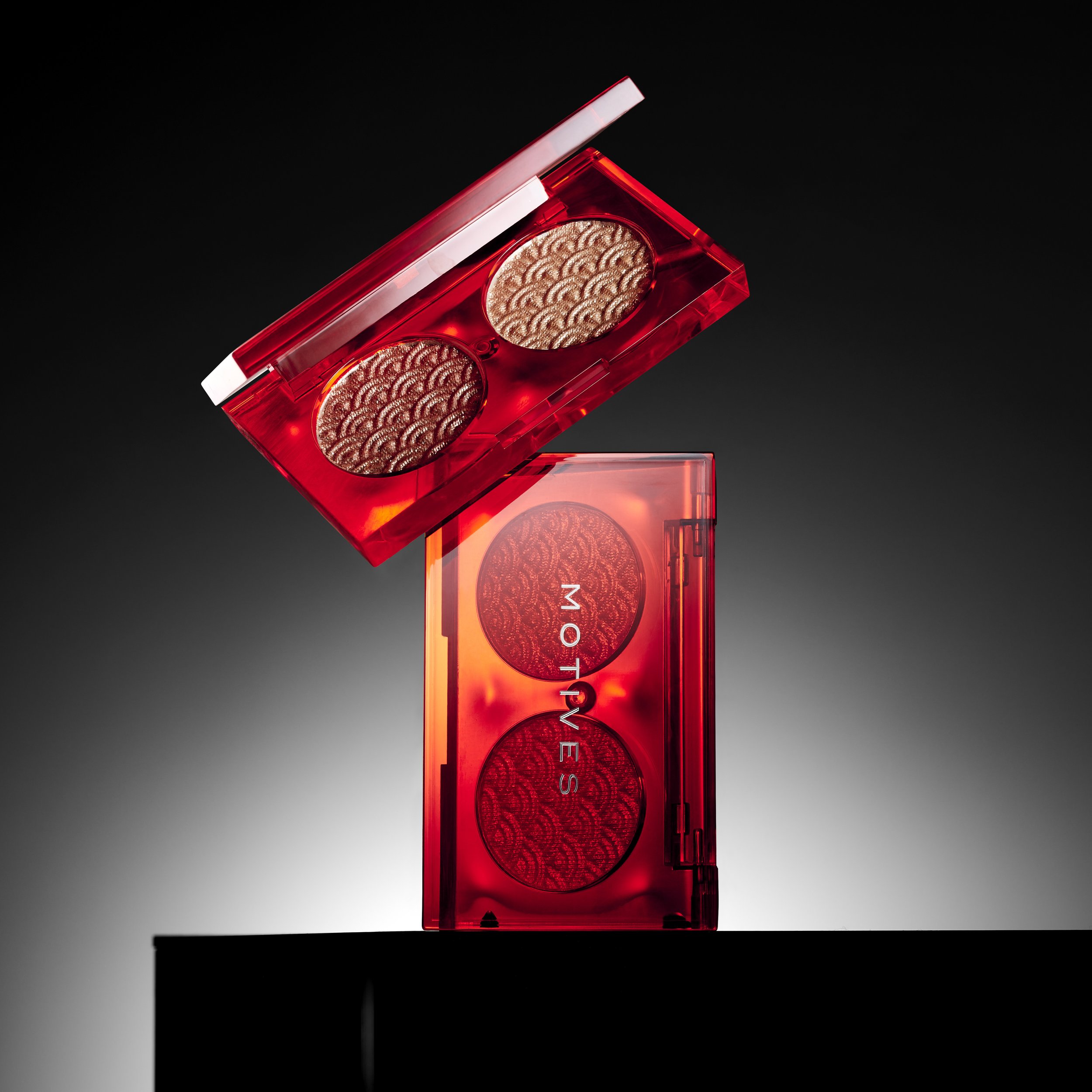 Red compact eyeshadow palette with three shimmery shades, one open to reveal two eyeshadows with wave-like patterns, and the other closed with the brand name 'MOTIVES' on it, placed against a gradient background.