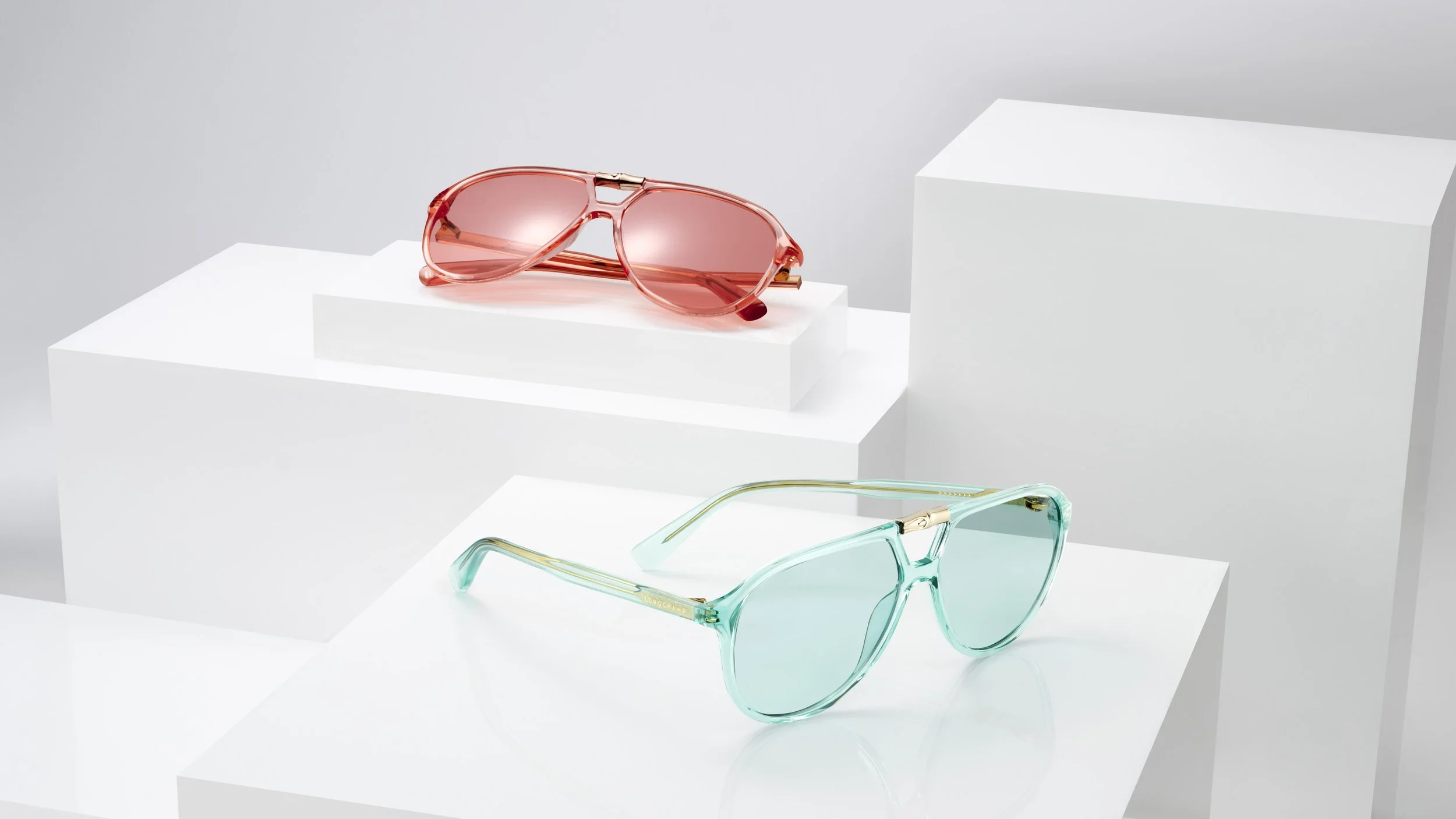 Two pairs of stylish sunglasses, one in pink with tinted lenses on a white display platform, and the other in turquoise with reflective lenses on a white surface, against a minimalist white background.