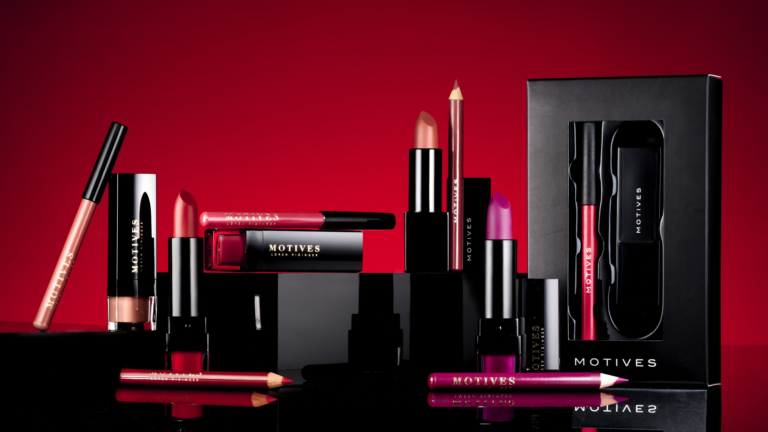 Collection of Motives lip makeup products including lip gloss, lipsticks, lip pencils, and a boxed set, displayed against a red background with reflections.