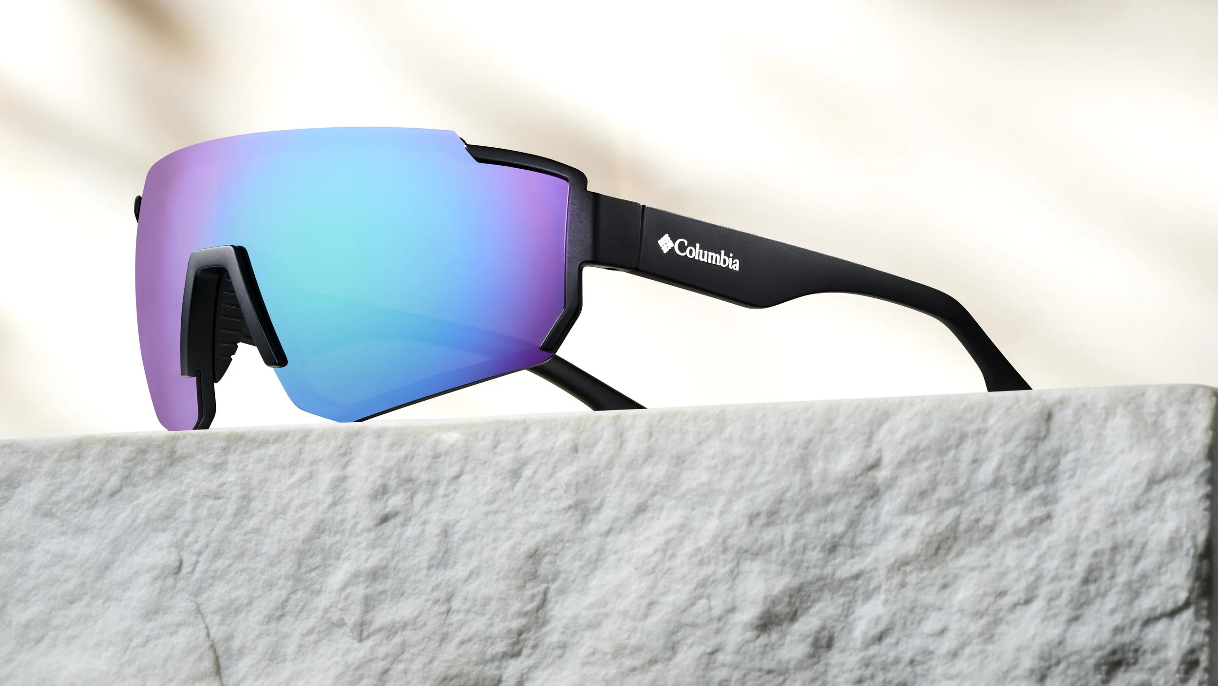 Black Columbia sports sunglasses with iridescent blue and purple lenses resting on a light-colored stone surface.