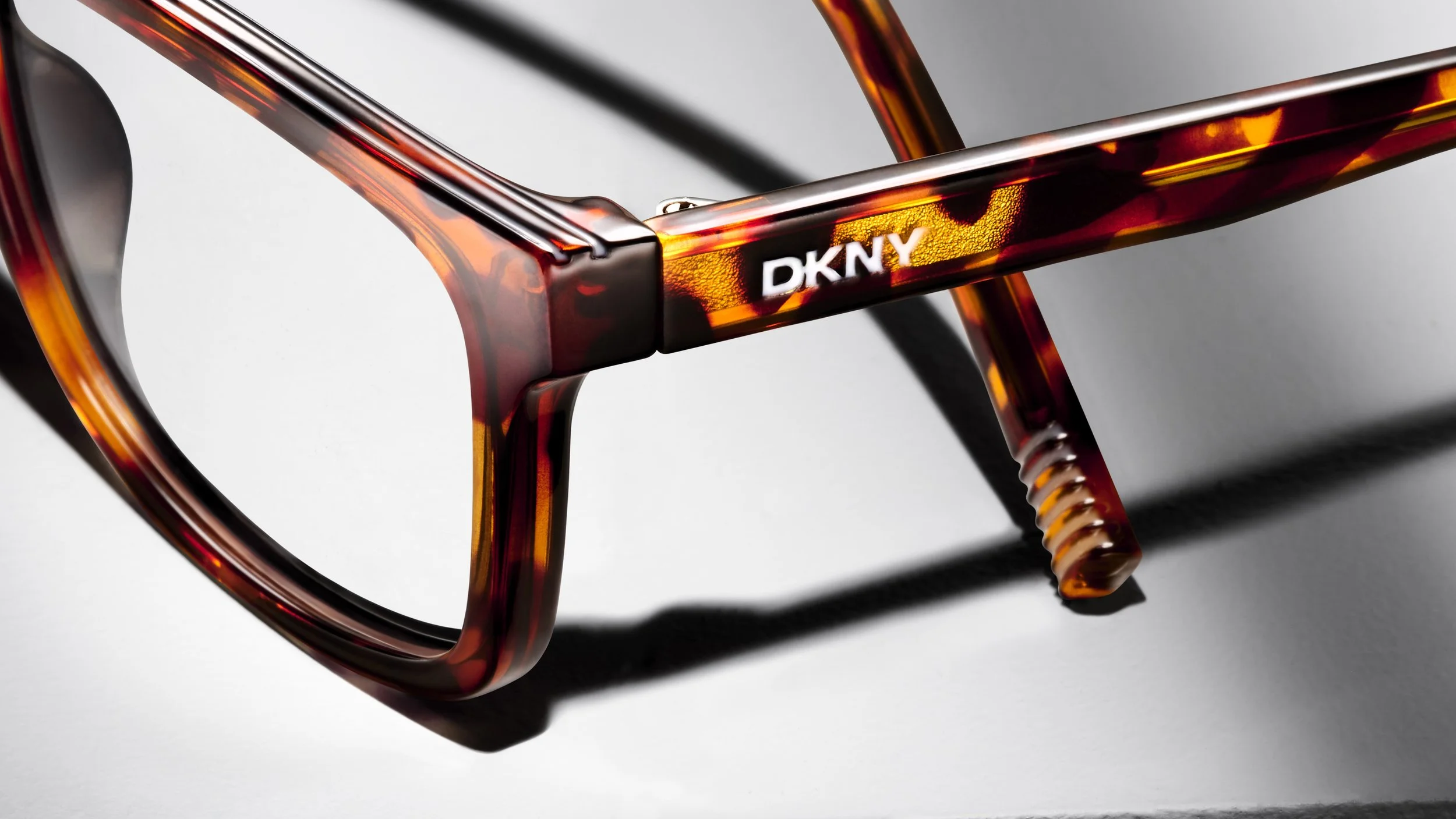 Close-up of tortoiseshell glasses with the brand DKNY printed on the inner side of the arm, casting a shadow on a flat surface.