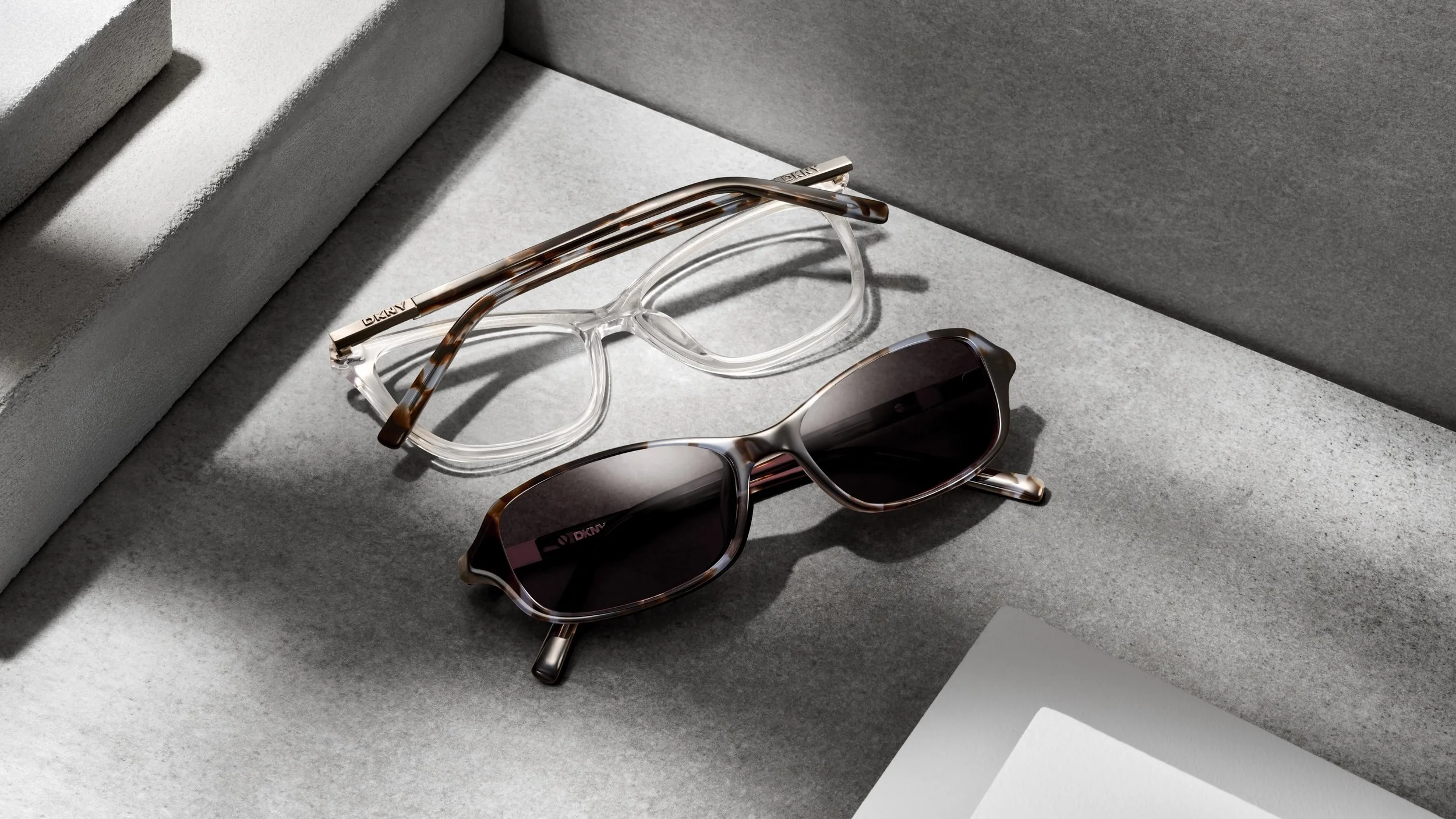 Two pairs of sunglasses, one with clear frame and the other with tortoiseshell frame, placed on a textured gray surface with shadows cast from overhead lighting.