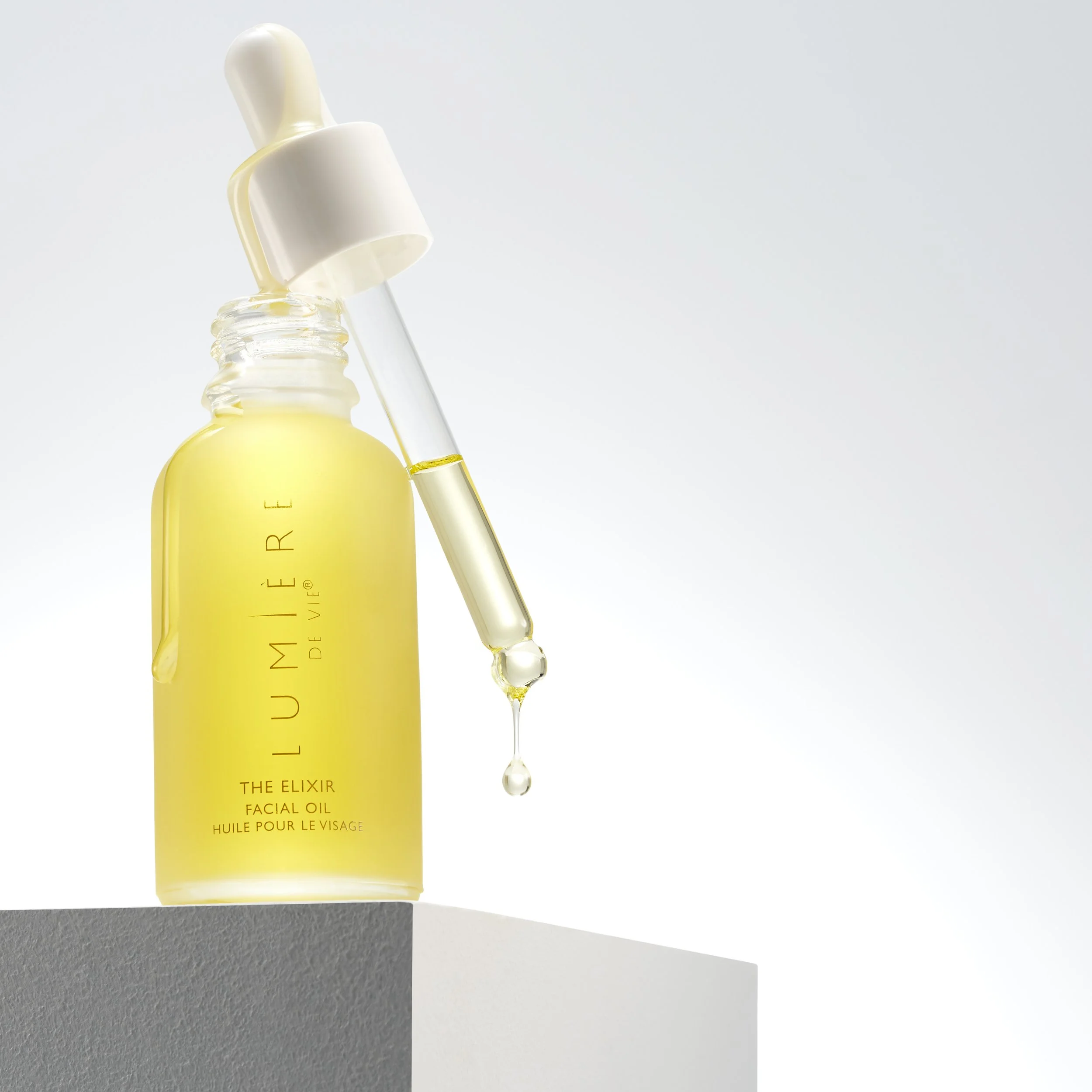 A yellow facial oil bottle with a dropper, with some facial oil and droplets hanging from the dropper, against a light gradient background.